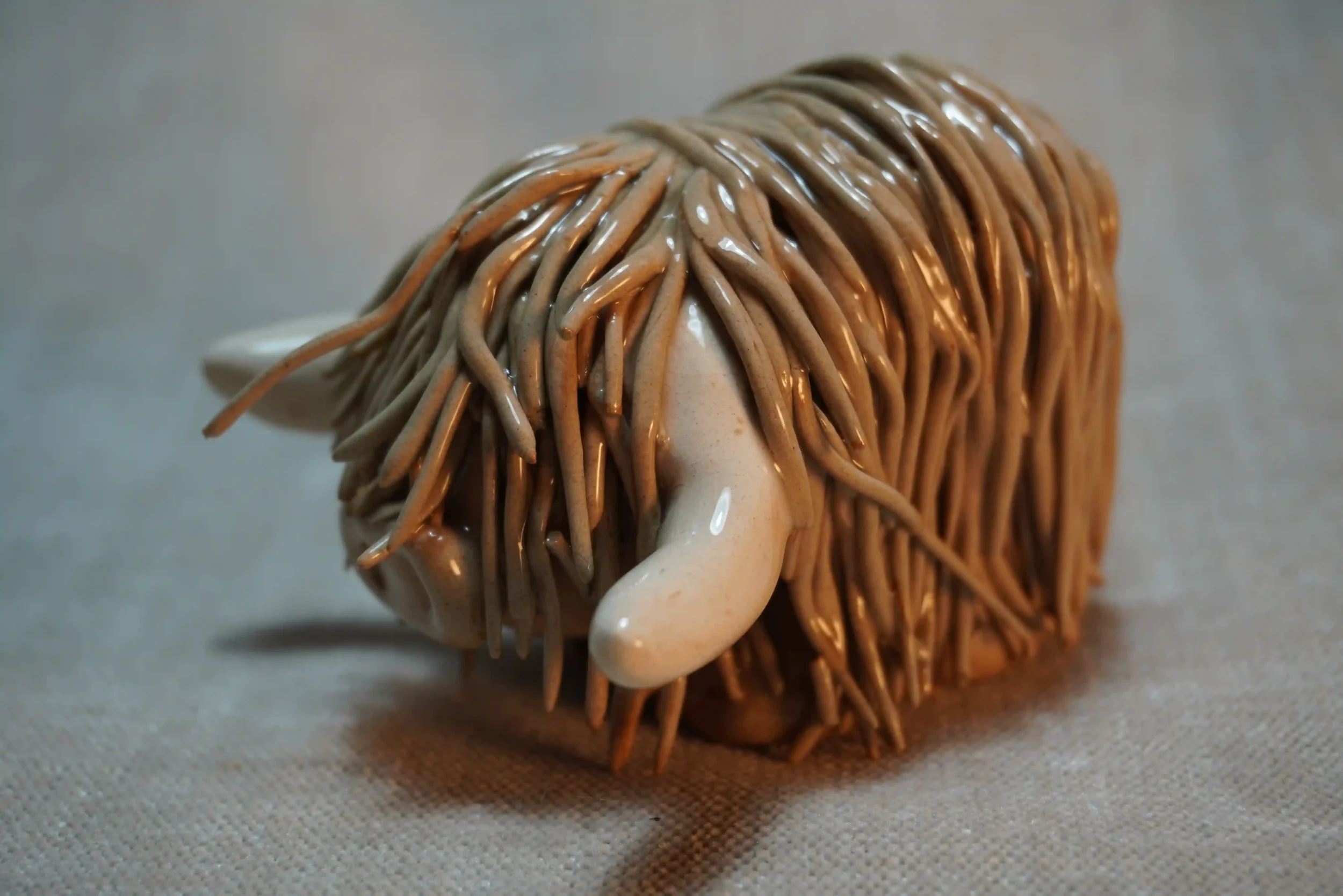 Side view of a handmade pottery ceramic sculpture of a Highland Cow ornament with light brown-coloured hair and beige horns.