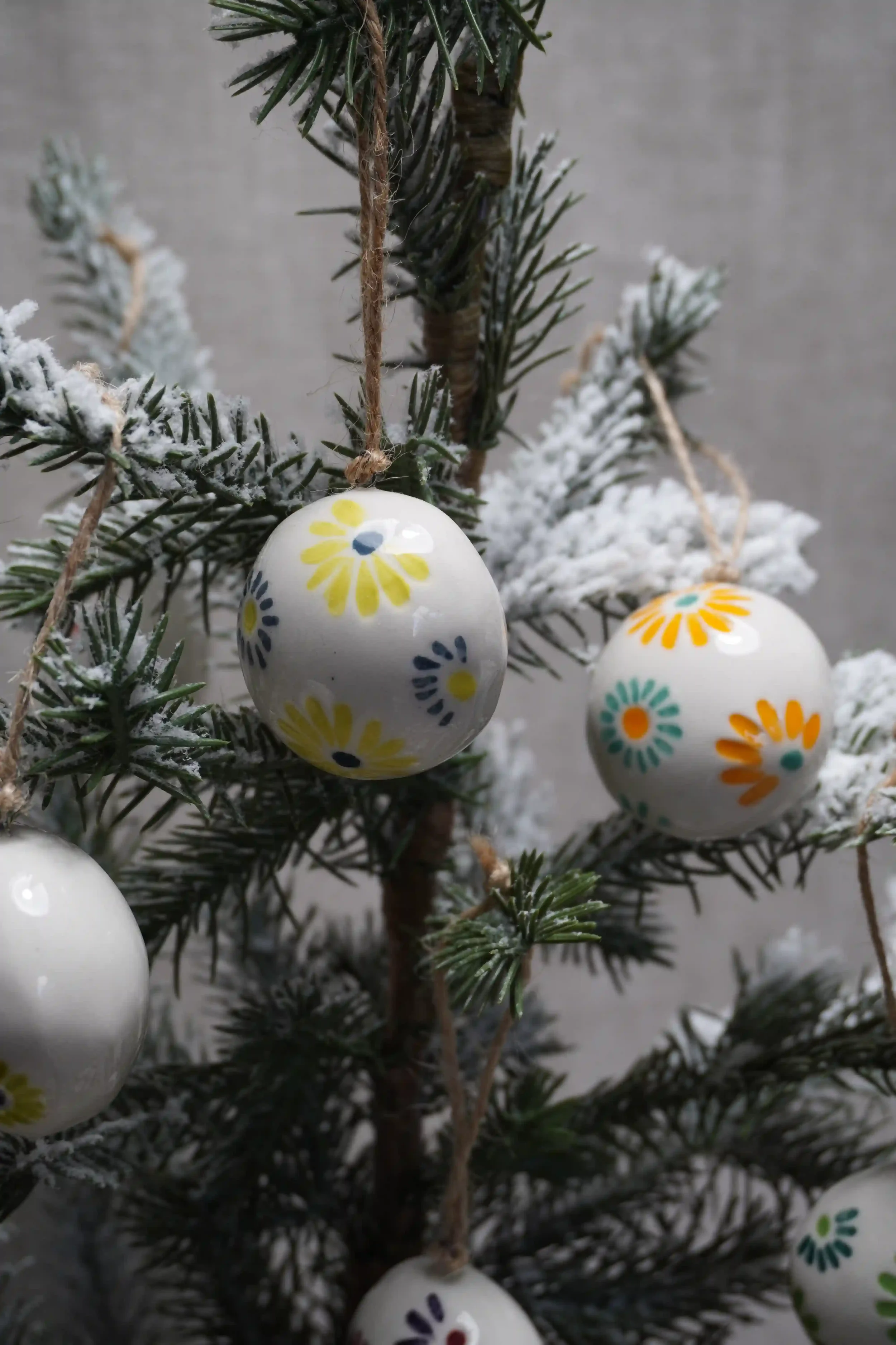 A close-up of a small Christmas tree decorated with white ceramic bauble ornaments featuring colorful firework patterns, with some branches having artificial snow.