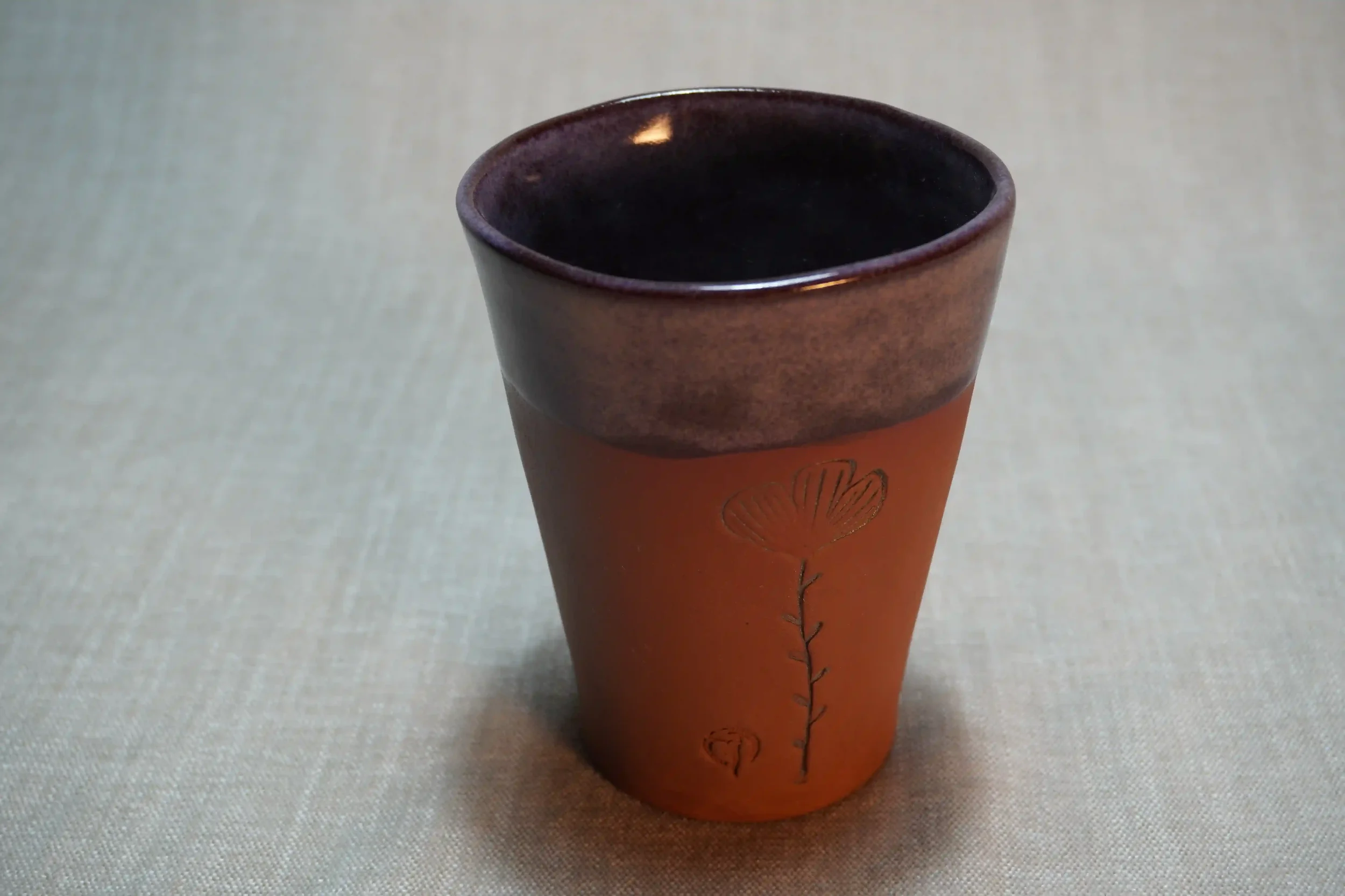 A ceramic pottery tumbler with purple glazed top and terracotta brown base and floral design
