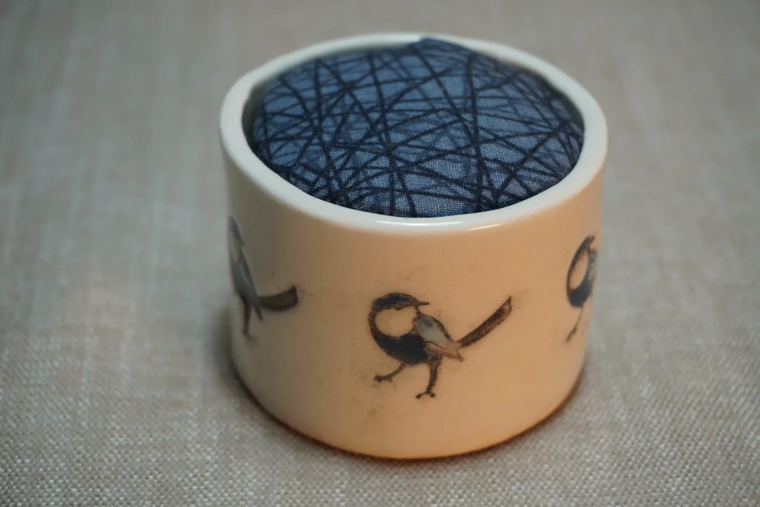 A ceramic pincushion with a blue bird design on the outside, filled with a patterned cloth cushion. The pincushion is shown on a textured surface.