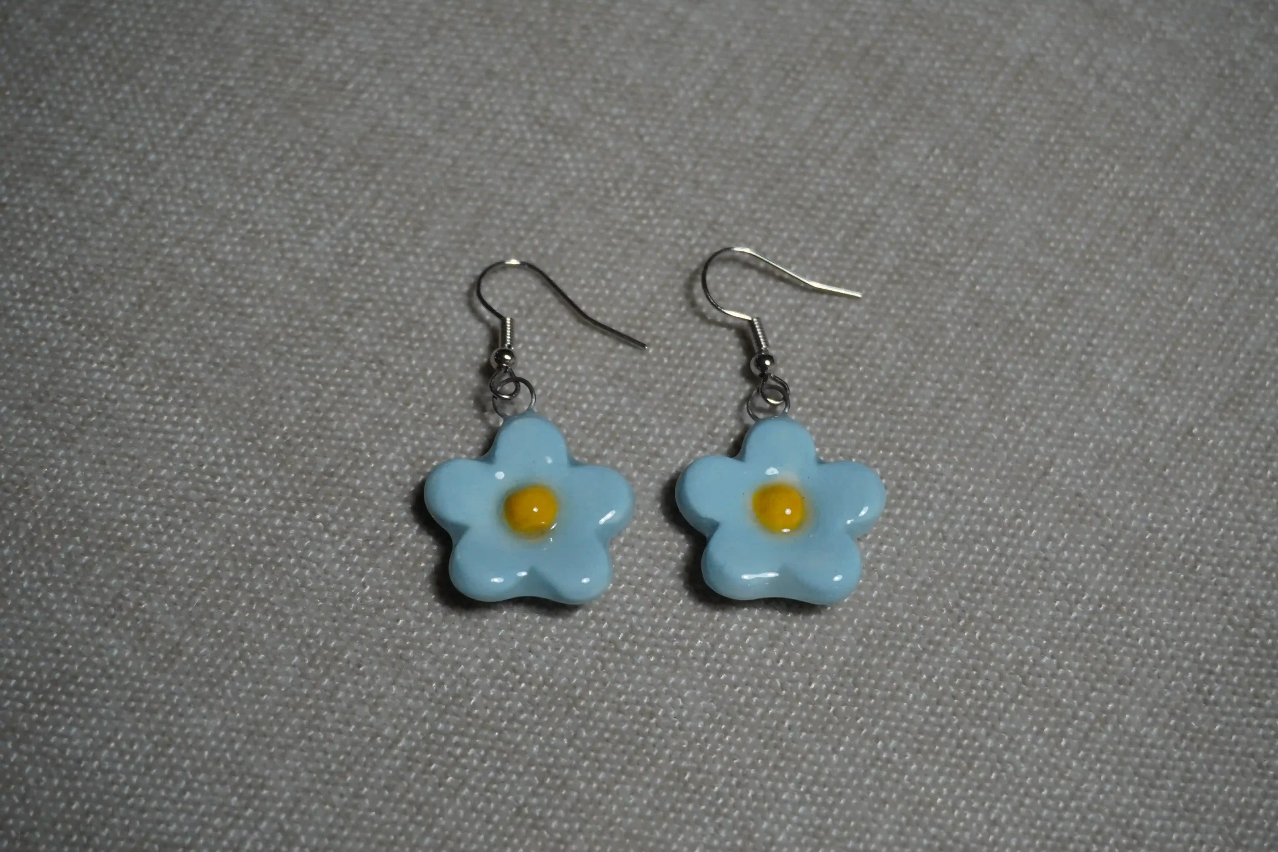 Pair of ceramic pottery earrings with blue flower-shaped pendants and yellow centers on a textured gray background.