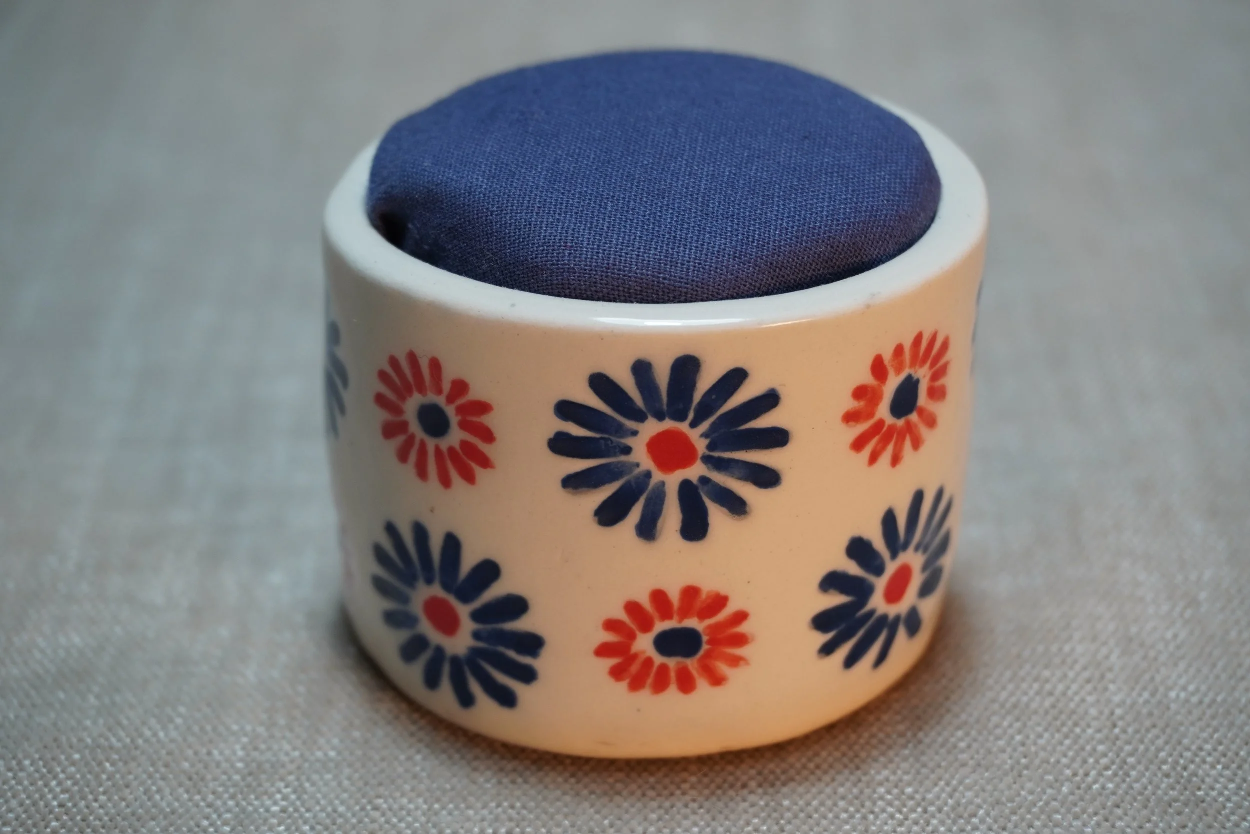 A ceramic pincushion with a red and blue firework pattern, filled with a blue cloth cushion. The pincushion is shown on a textured surface.