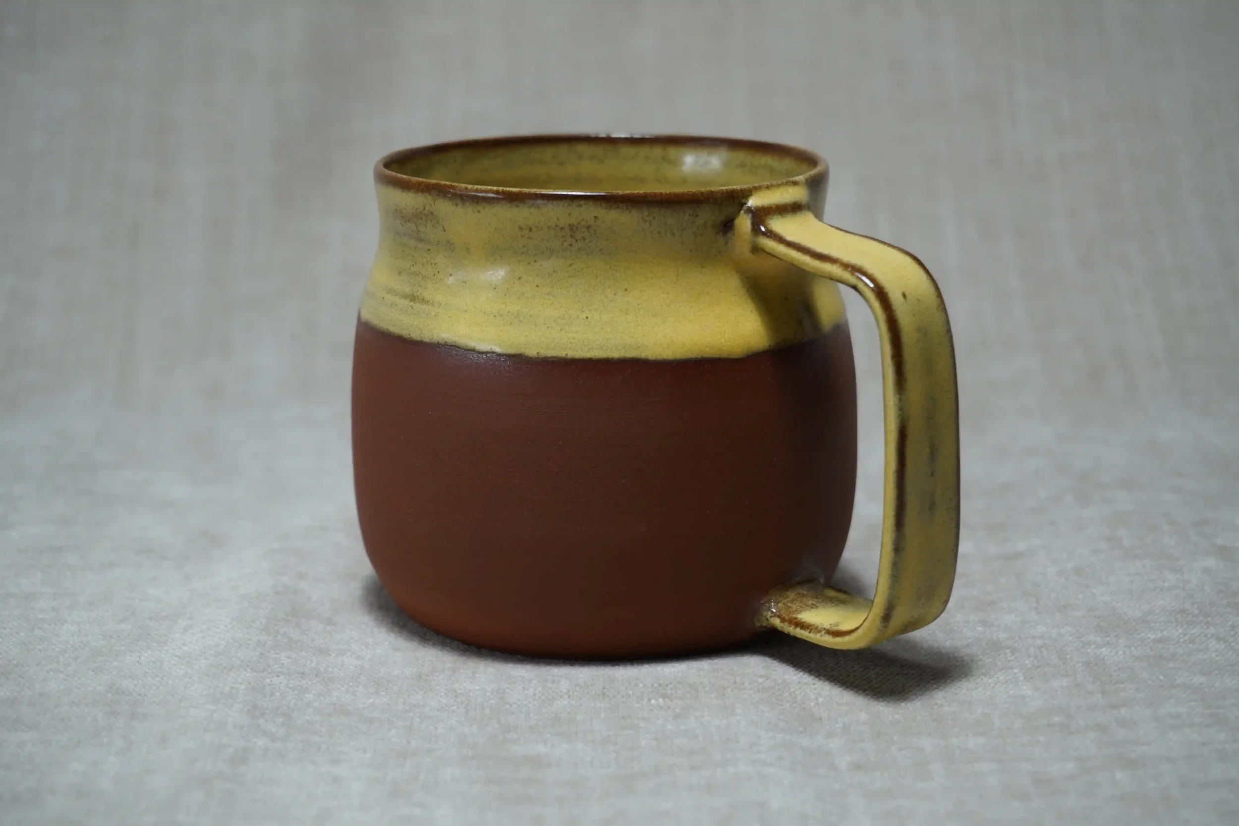 A ceramic mug with a yellow glazed top and an unglazed terracotta brown bottom, featuring a distinctive handle, placed on a light-colored surface.