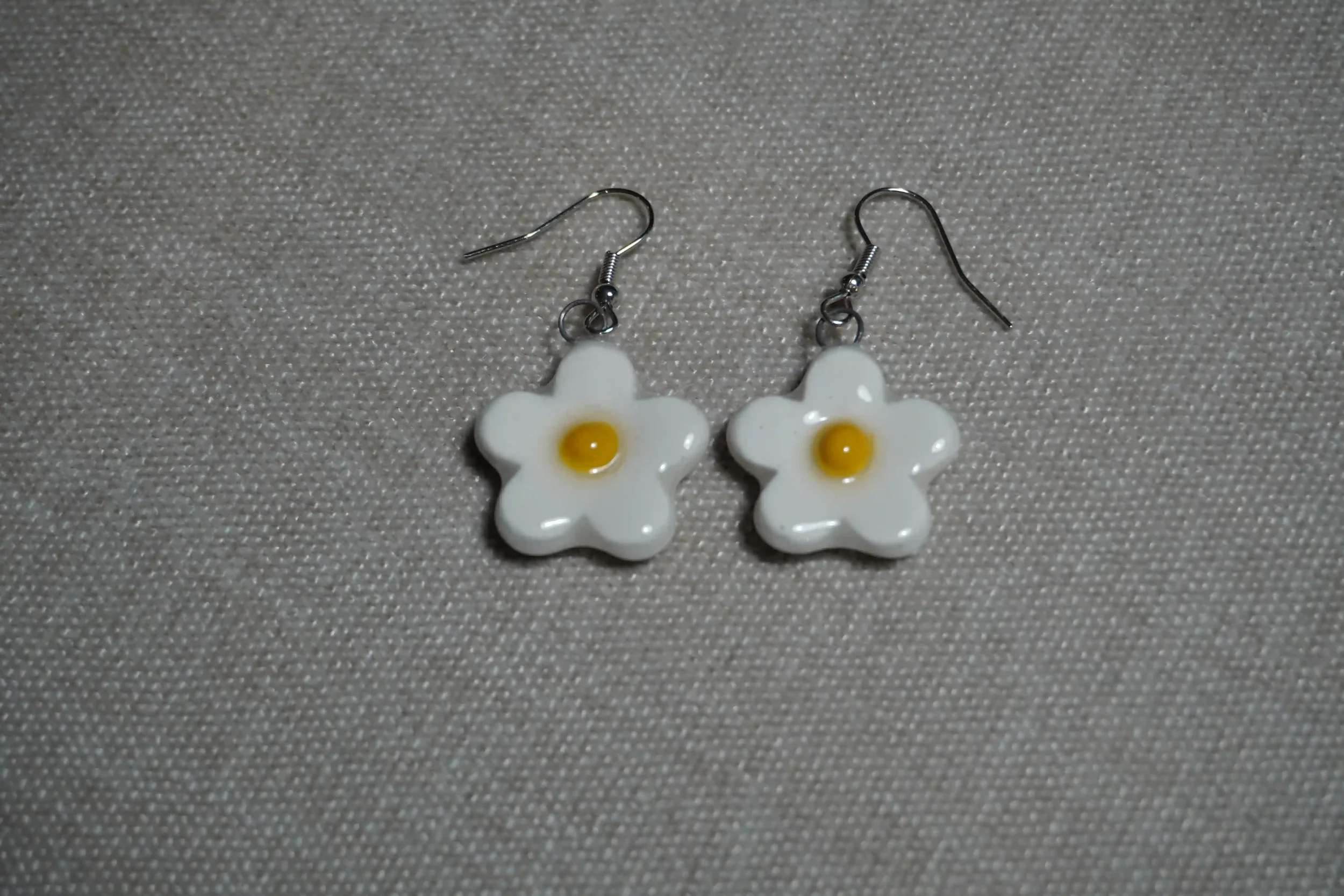 Pair of ceramic pottery earrings featuring white flower-shaped pendants with yellow centers, placed on a textured beige surface.