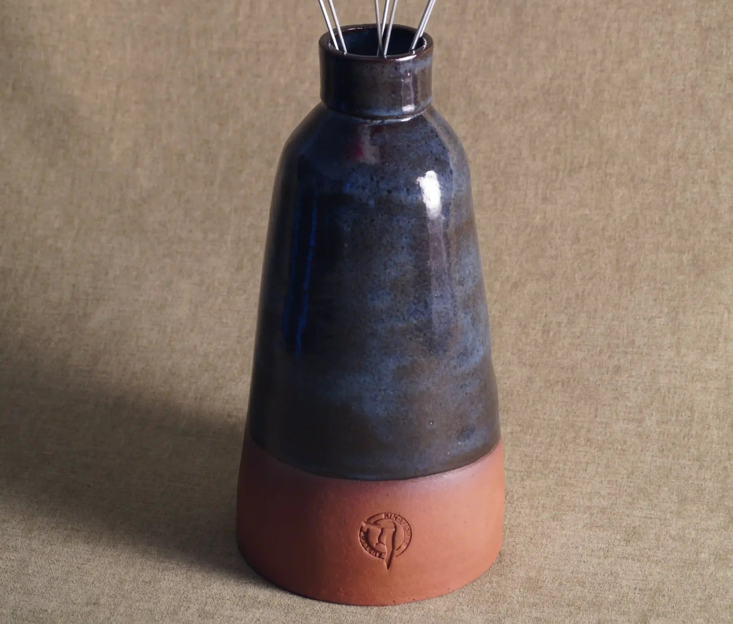 A ceramic vase with a wide, rounded terracotta base, narrow neck, and a blue glaze, positioned on a textured beige background.
