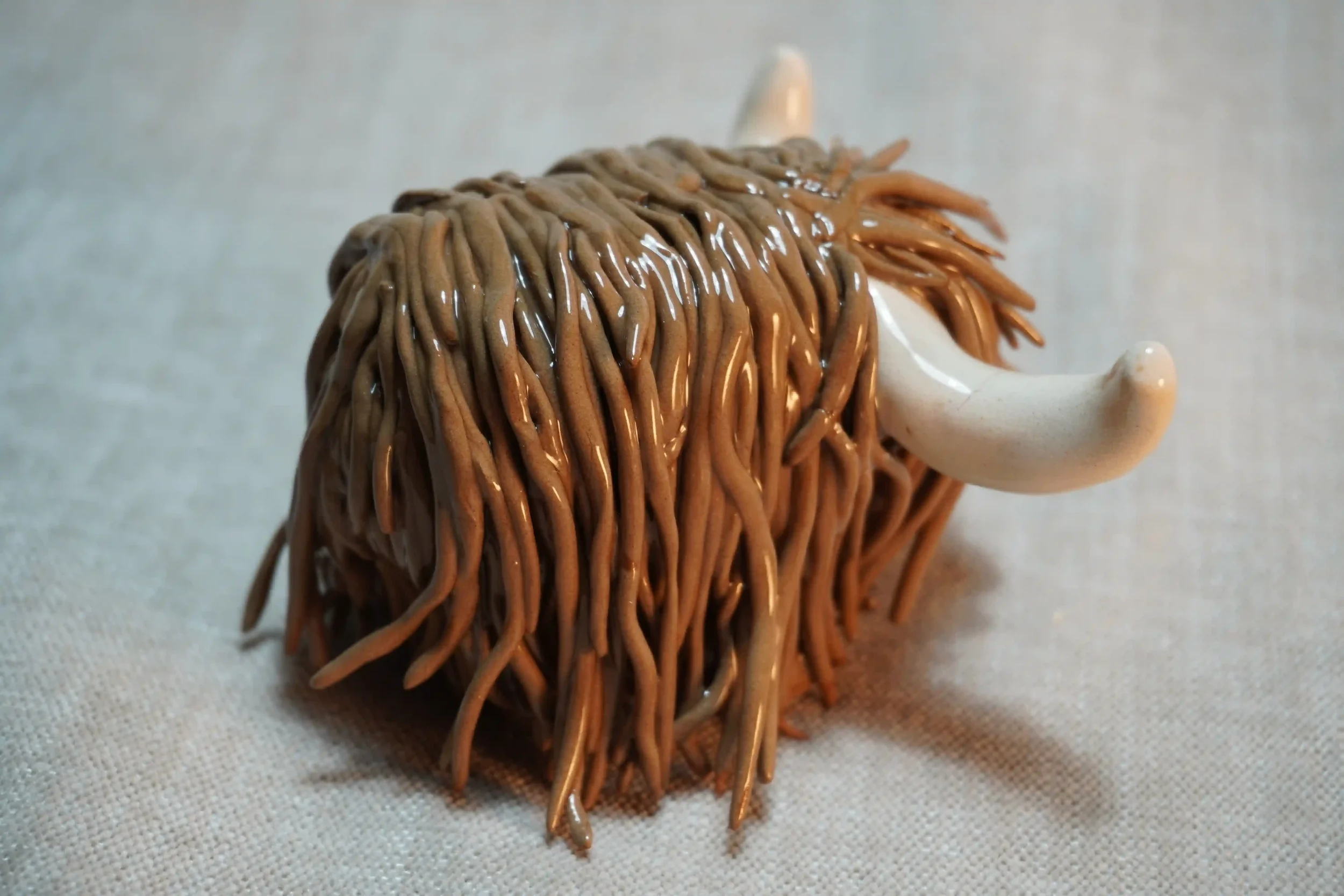 Side view of a handmade pottery ceramic sculpture of a Highland Cow ornament with milk chocolate-coloured hair and beige horns.