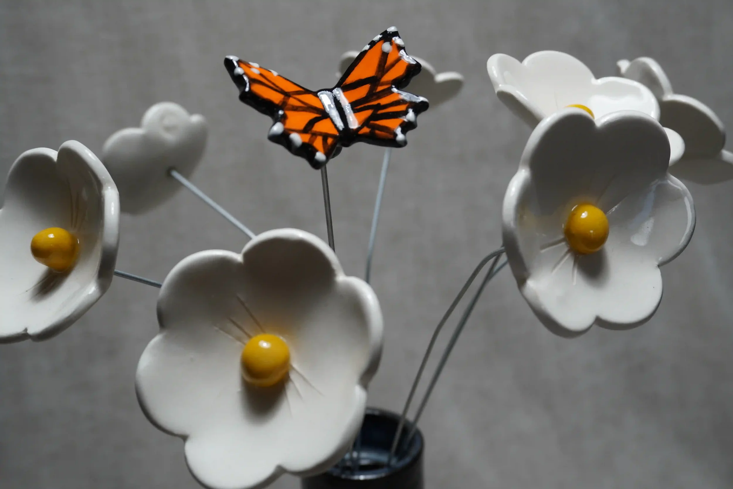 White handmade ceramic Daisy flowers with yellow centers, and a ceramic Monarch butterfly with orange and black wings, arranged among the flowers.
