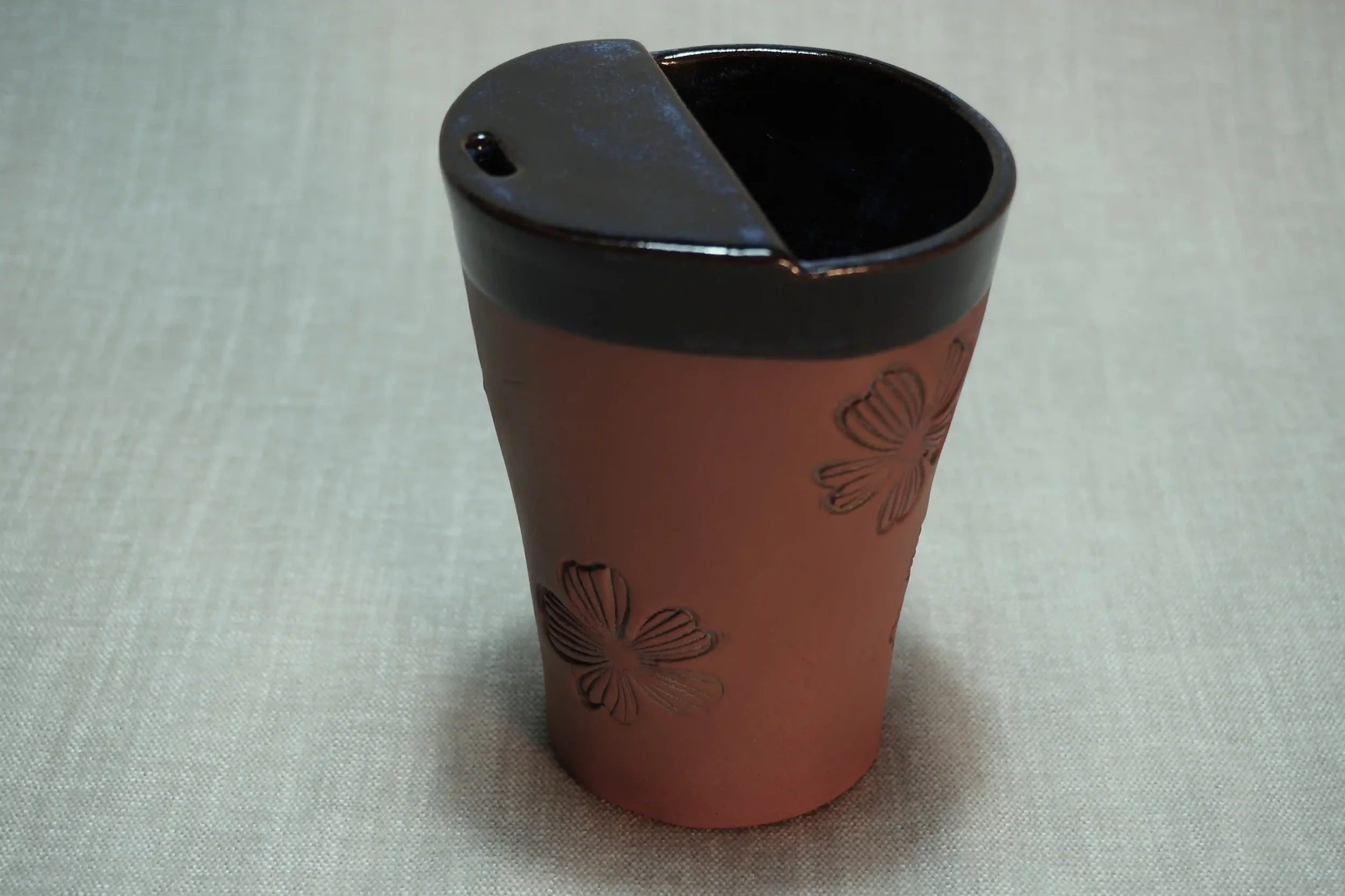 A ceramic travel mug with a dark blue glaze, a terracotta brown base, and a decorative floral design on the outside, placed on a light-colored surface.
