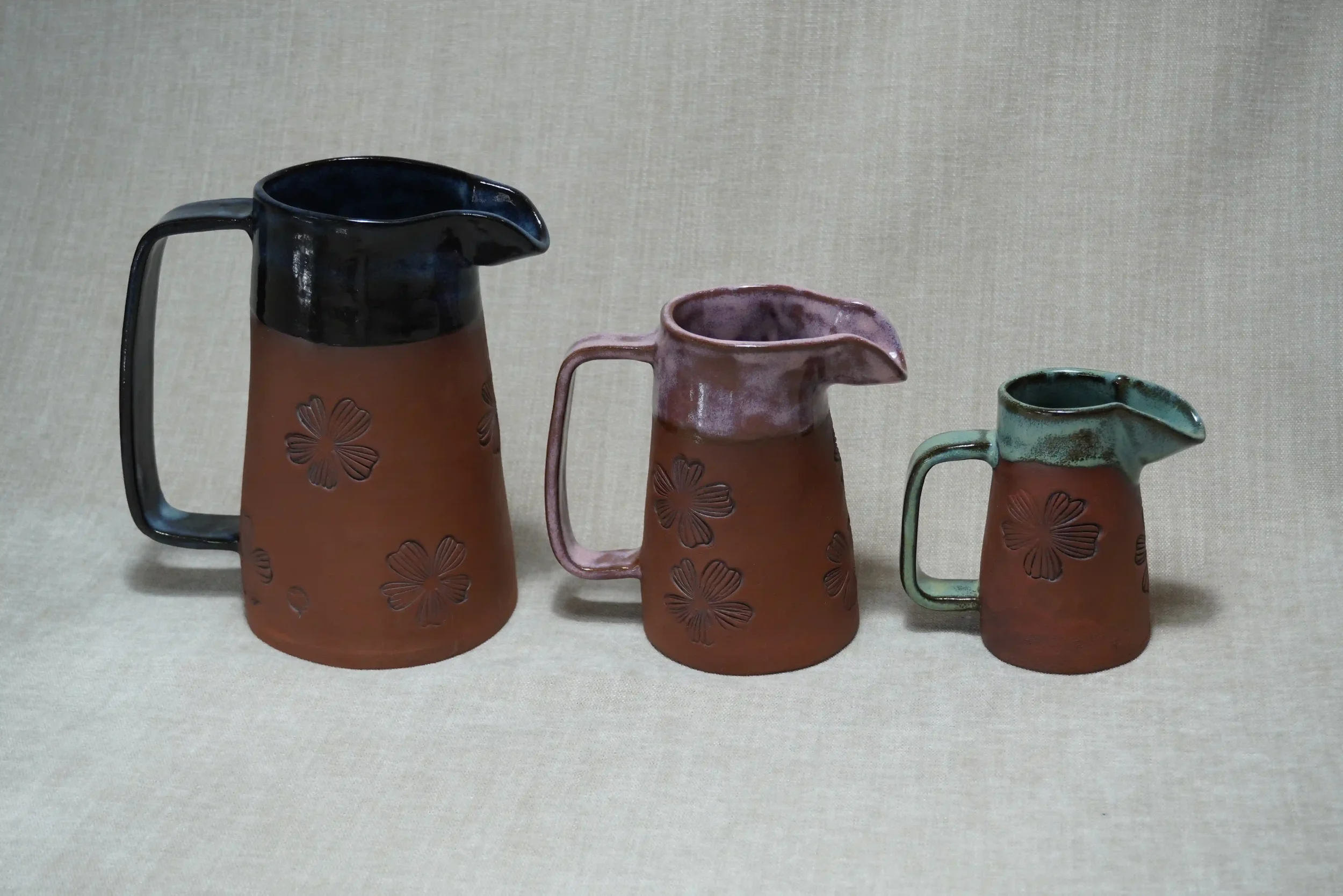 Three ceramic pitchers with floral patterns, each differing in size and color, arranged in a row against a plain background.