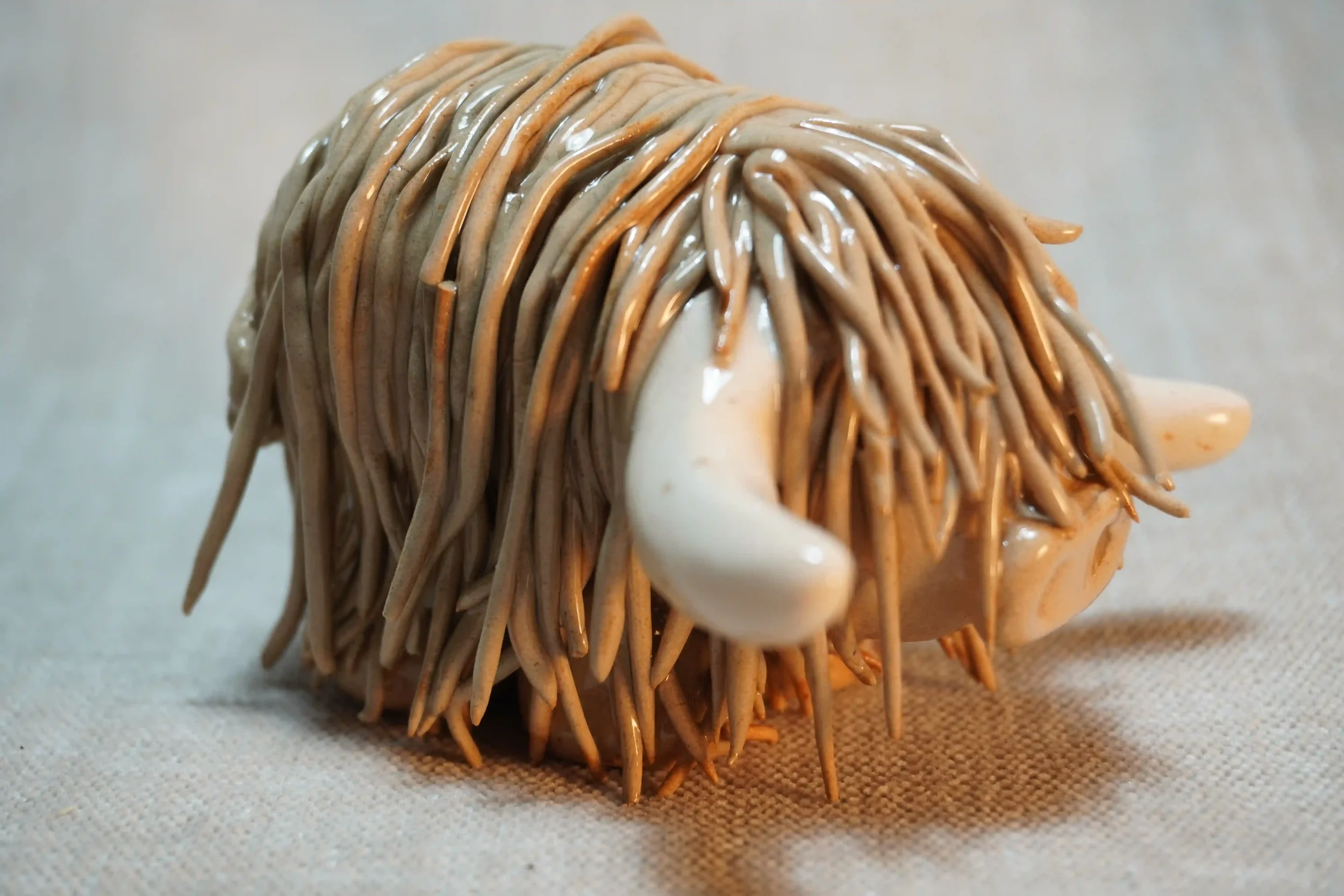 Side view of a handmade pottery ceramic sculpture of a Highland Cow ornament with light brown-coloured hair and beige horns.