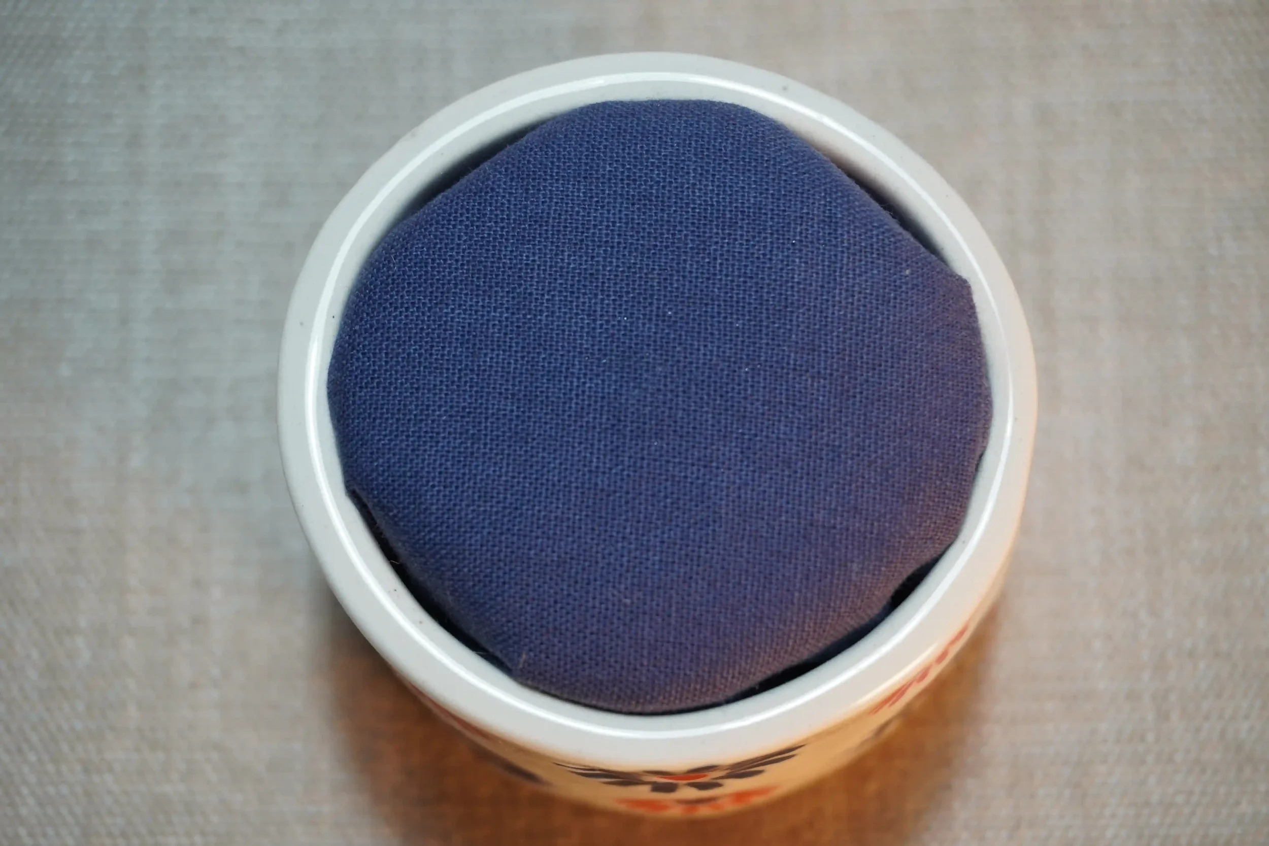 View from above of a ceramic pincushion with a blue cloth-covered cushion. The pincushion is shown on a textured surface.