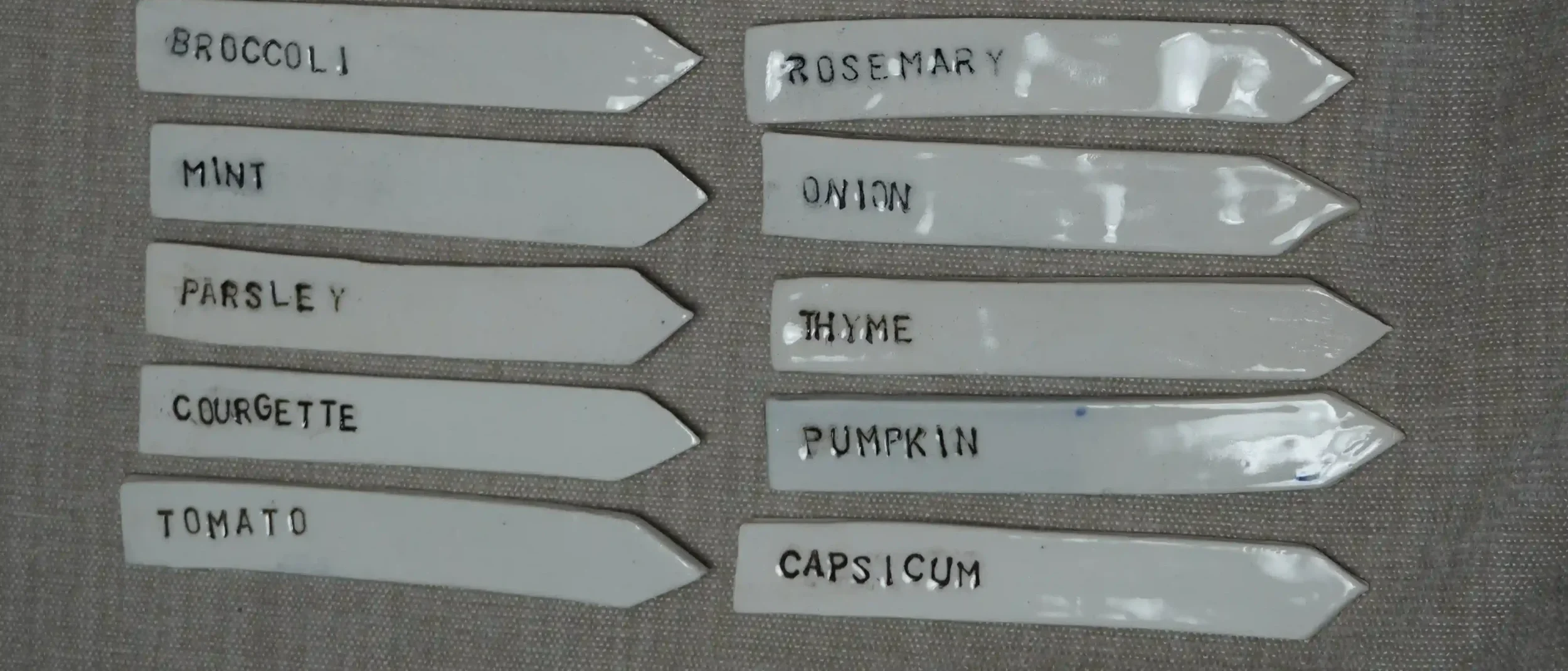 White ceramic plant labels with black text for gardening plants: broccoli, rosemary, mint, onion, parsley, thyme, courgette, pumpkin, tomato, capsicum.