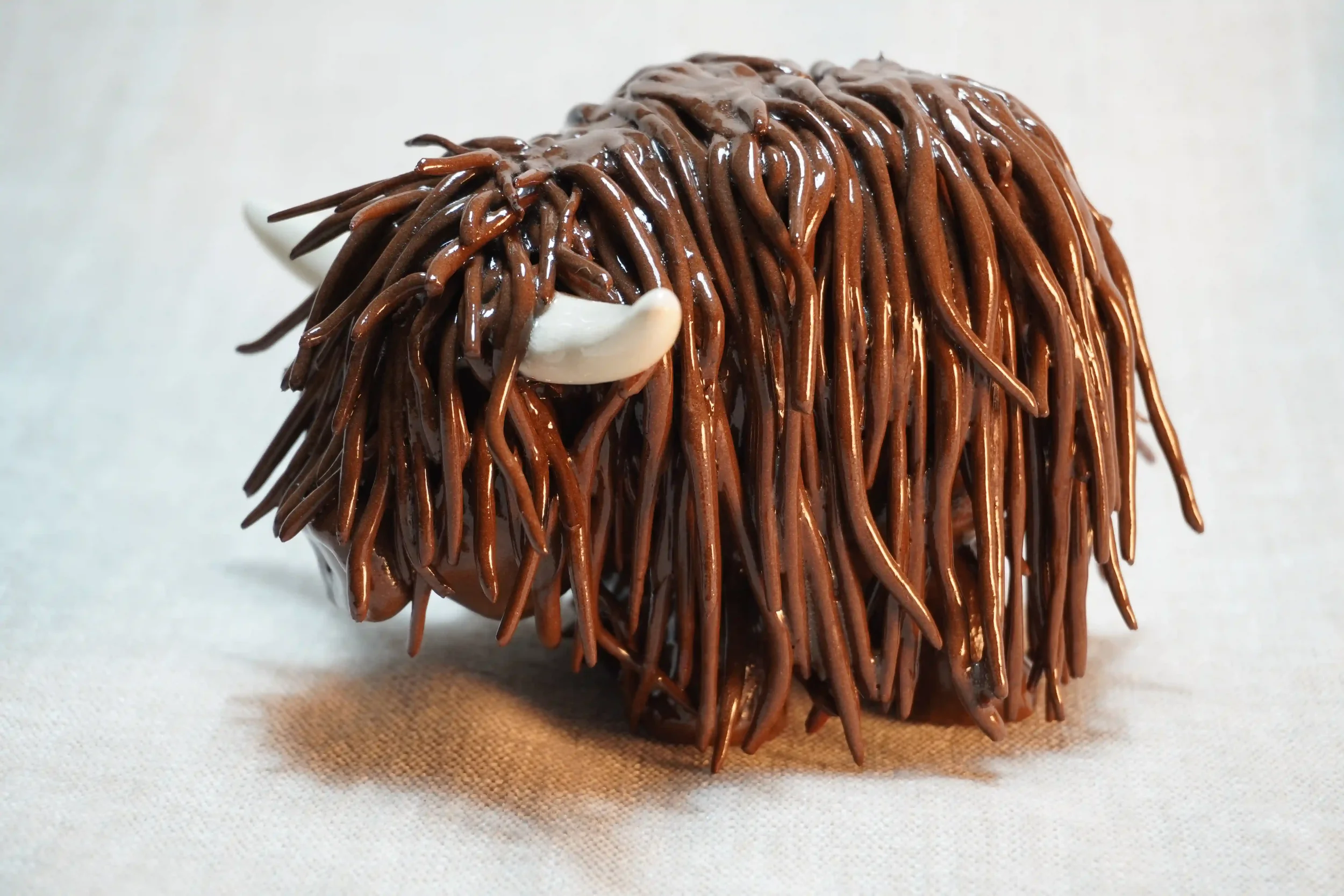 	
Side view of a handmade pottery ceramic sculpture of a Highland Cow ornament with deep brown-coloured hair and beige horns.
