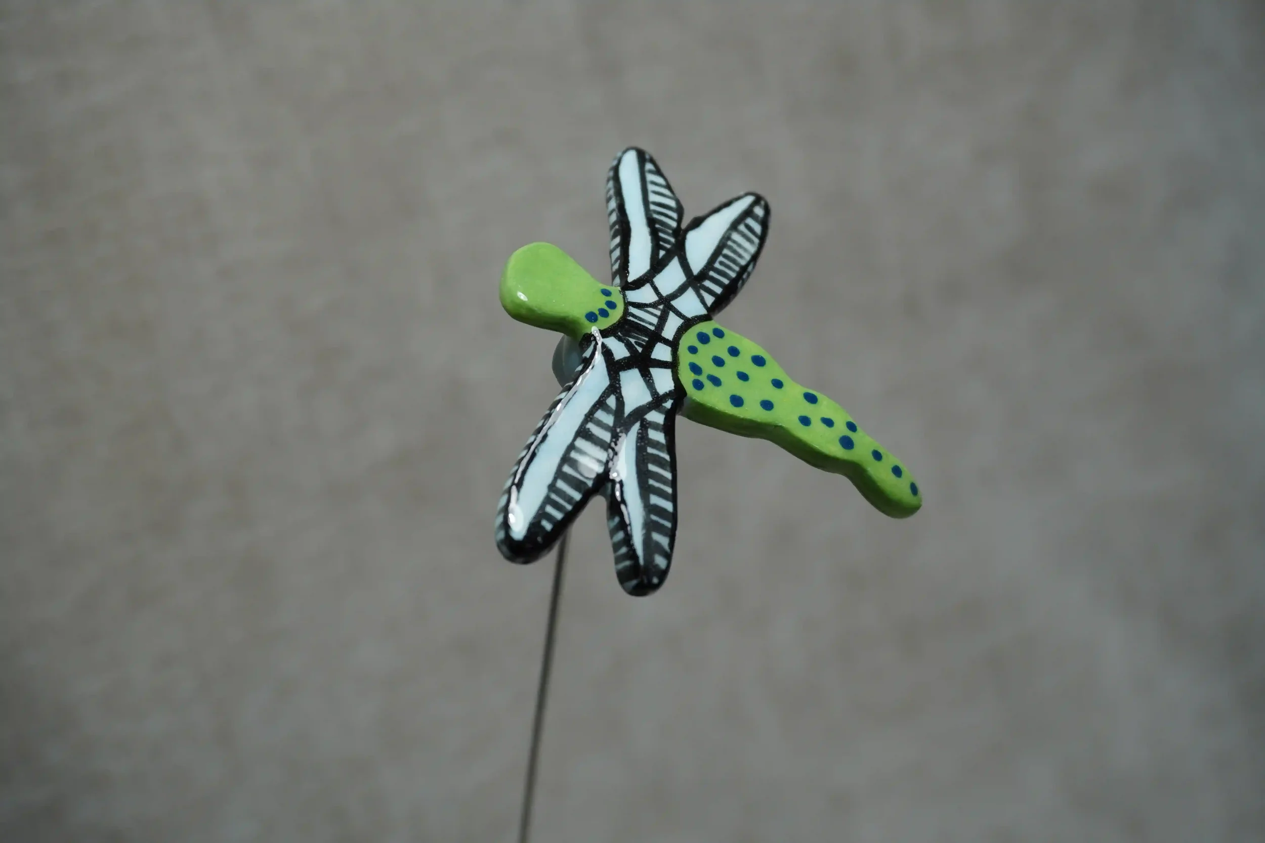 A ceramic pottery dragonfly with a green body with blue specks and light blue and black wings on a metal stem.