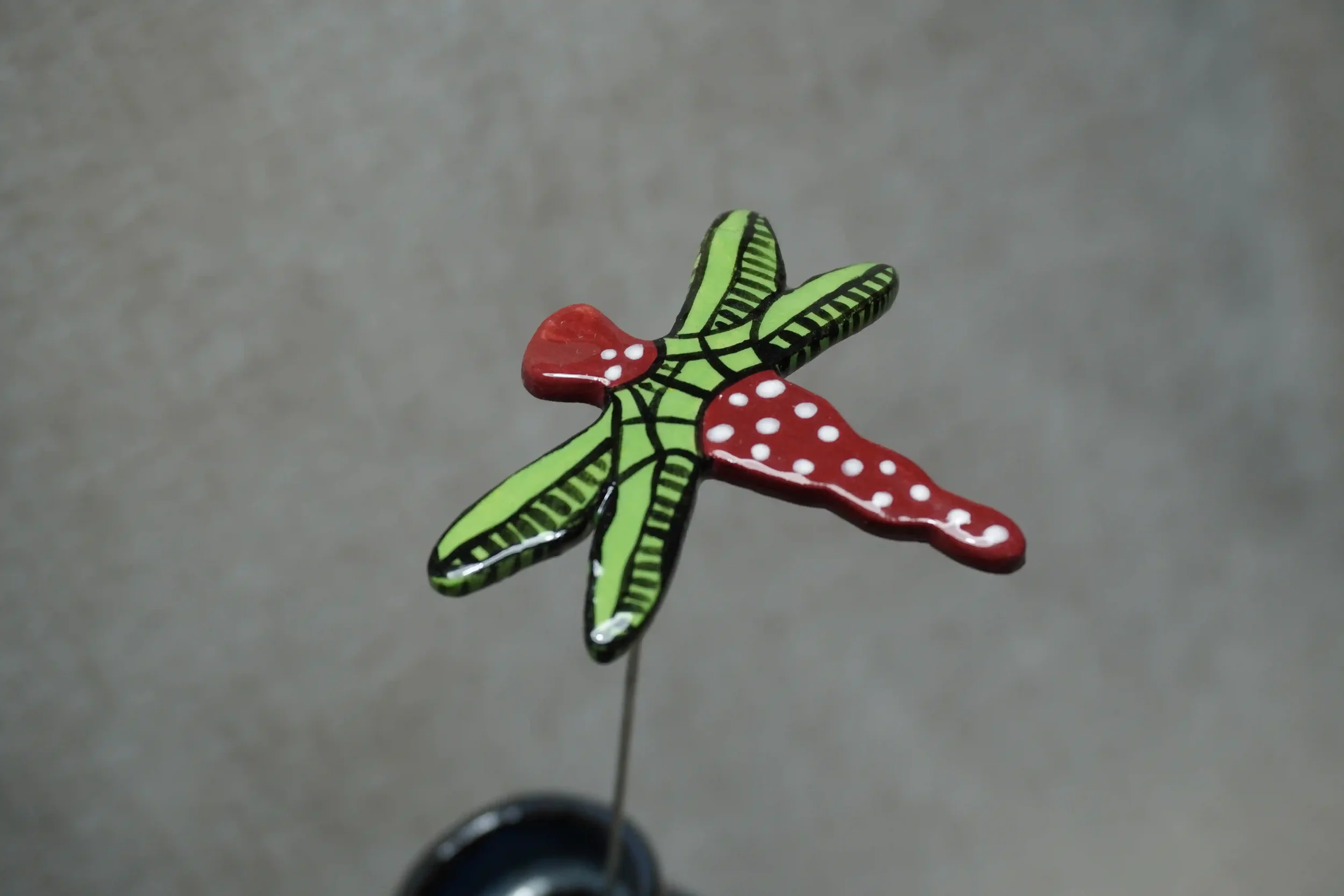 A handmade ceramic pottery dragonfly with red body with white speckles and green and black wings. Mounted on a metal stem.