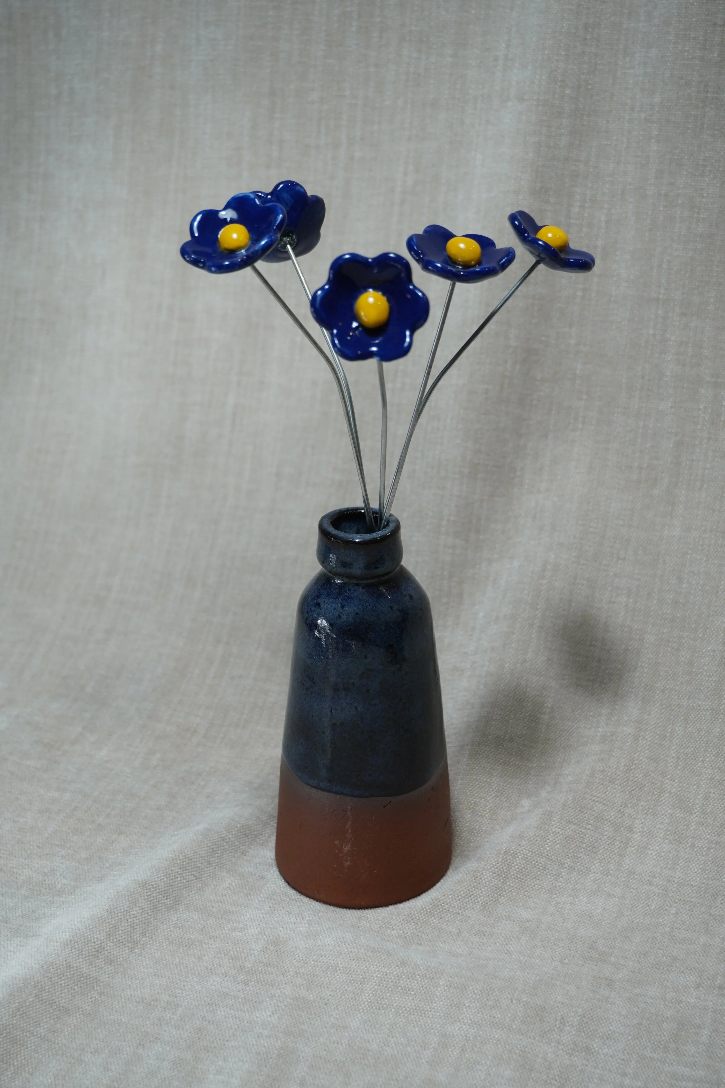A ceramic vase with a wide, rounded terracotta base, narrow neck, and a blue glaze, holding five blue ceramic flowers with yellow centers and metal stems, set against a neutral fabric background.