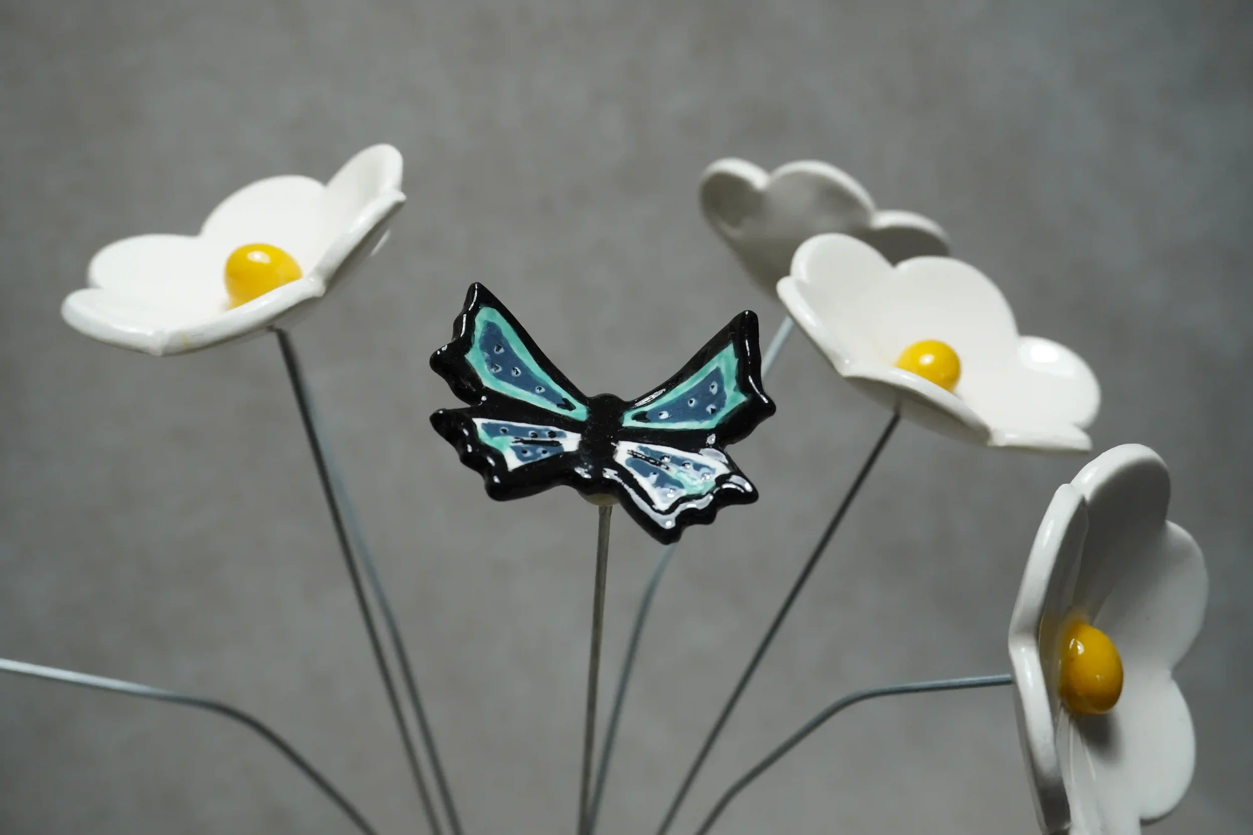 Handmade ceramic decorative white Daisy flowers with yellow centres, and a blue, green and black glazed butterfly on metal stems. 