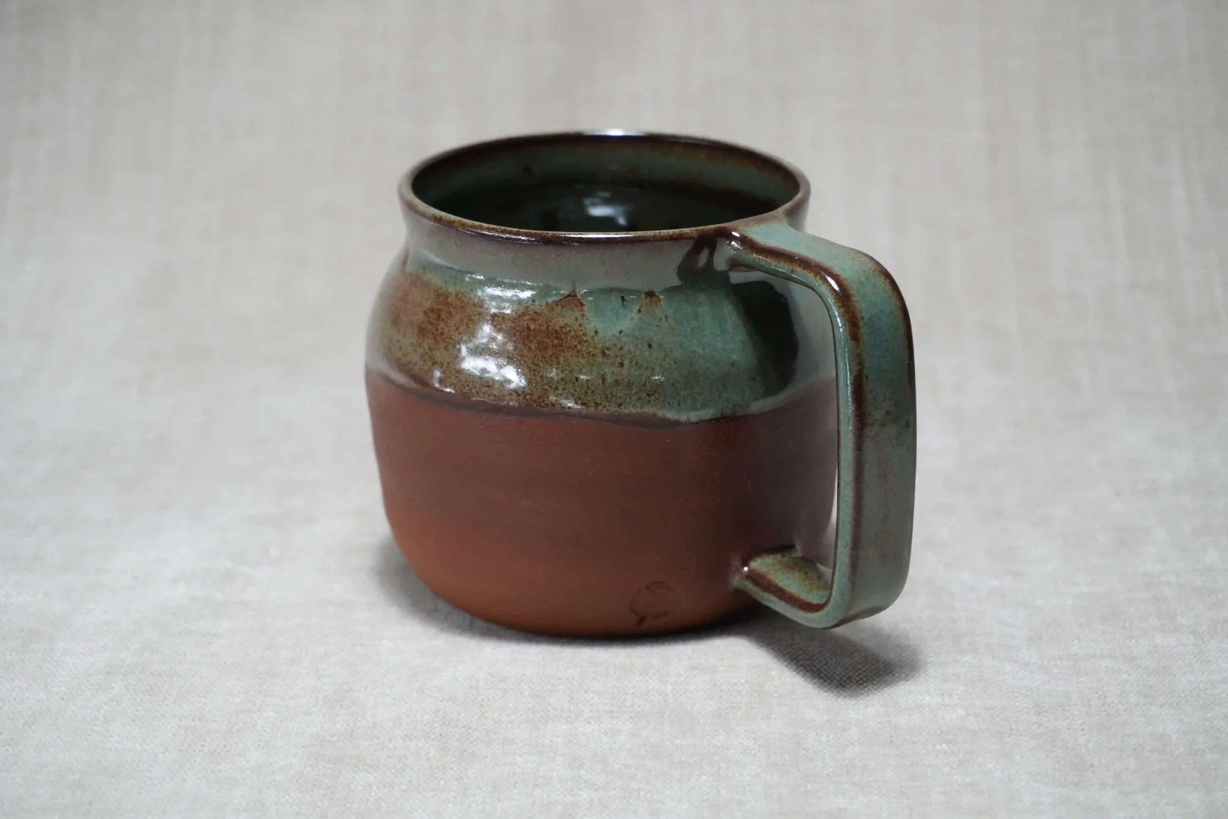 A ceramic belly mug with a green and brown glaze, with a matte terracotta brown bottom, displayed against a plain background.