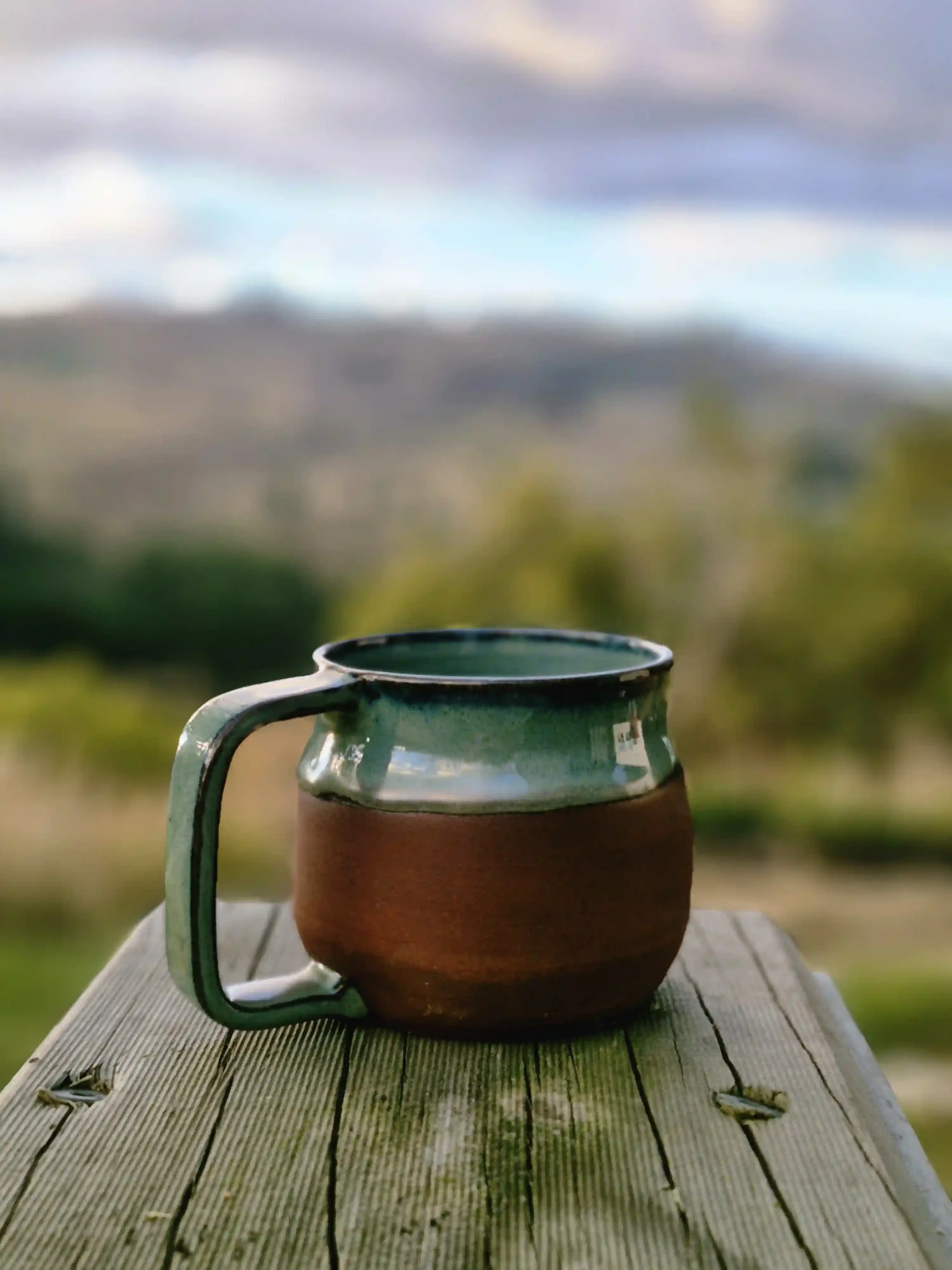 A close-up of a small ceramic belly mug with a handle, placed on a weathered wooden surface outdoors, with a blurred landscape of grass, trees, and a cloudy sky in the background.
