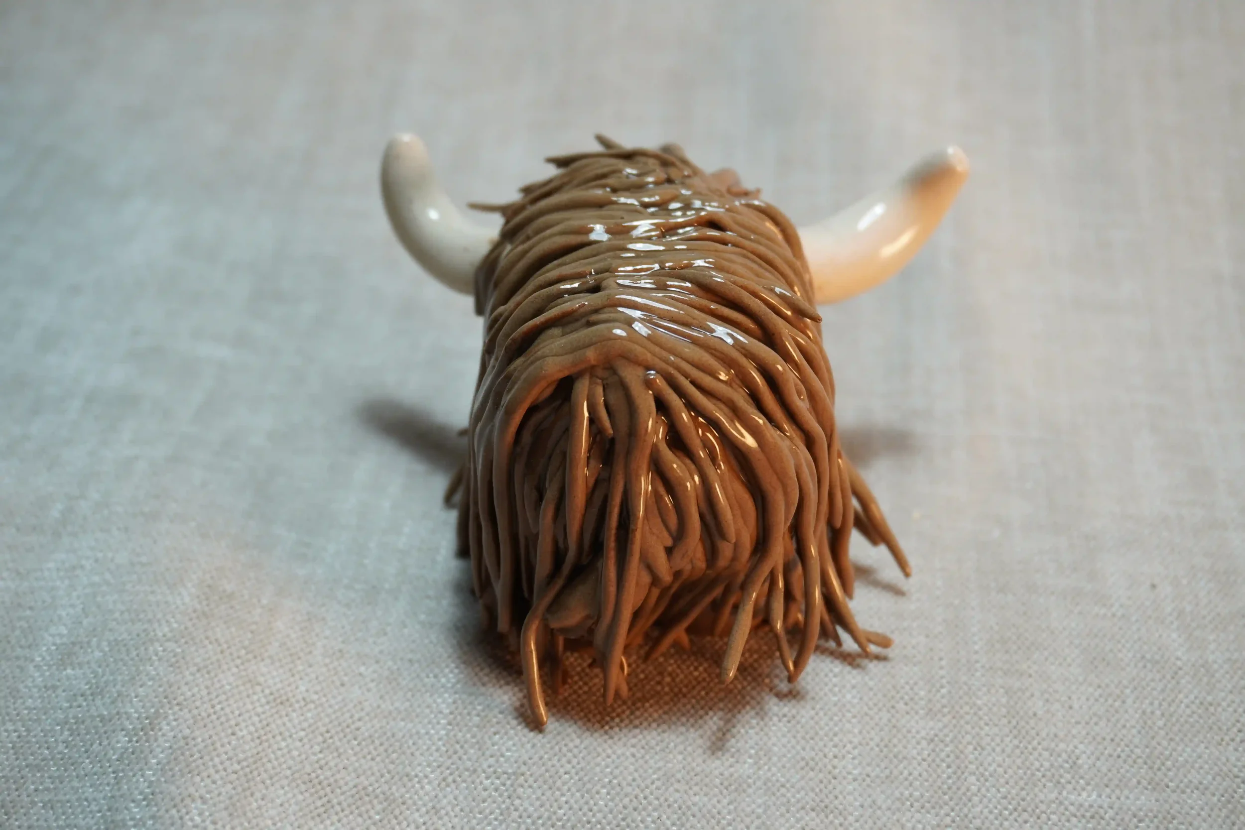 Rear view of a handmade pottery ceramic sculpture of a Highland Cow ornament with milk chocolate-coloured hair and beige horns.