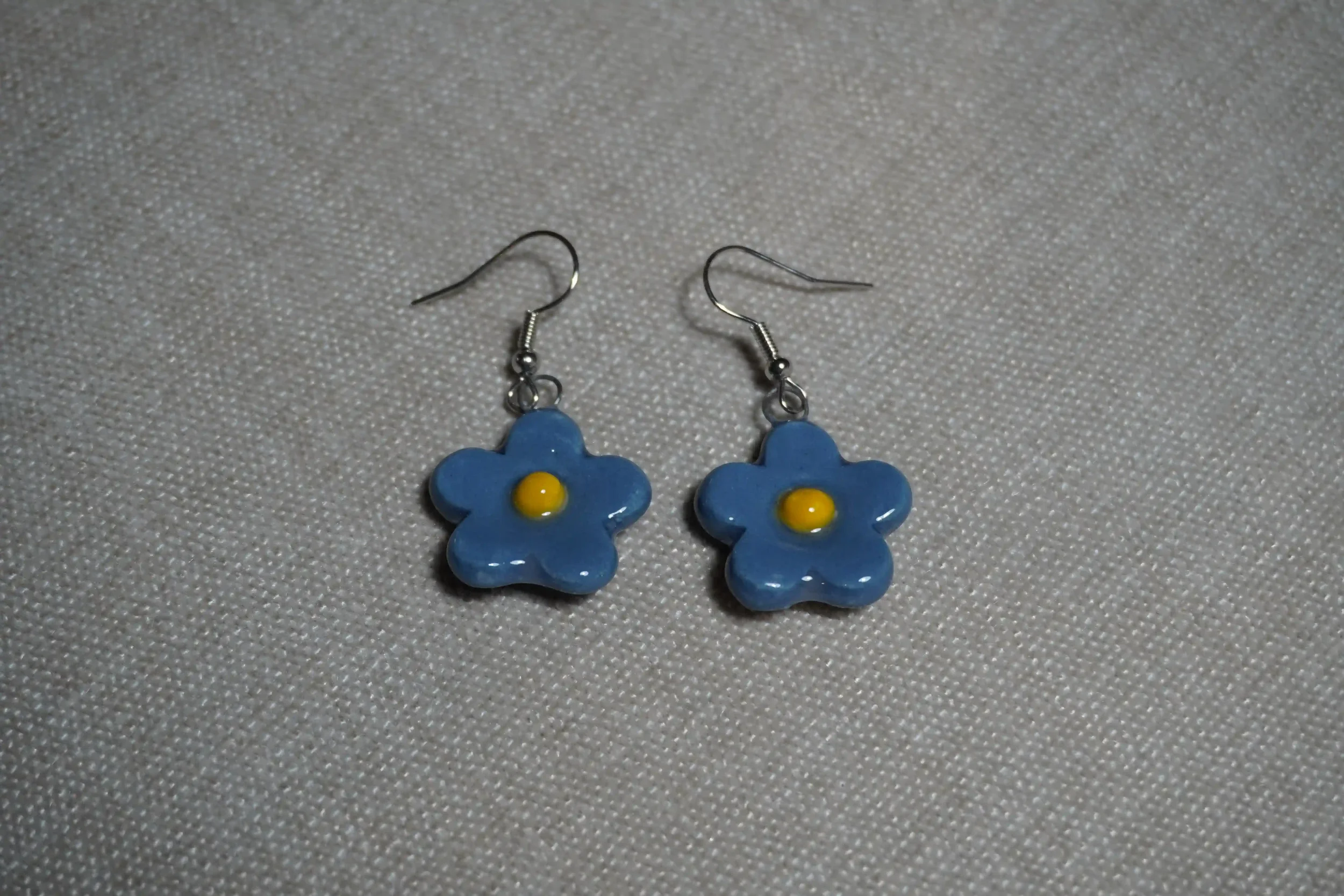 A pair of ceramic pottery blue flower-shaped earrings with yellow centers on a textured gray background.