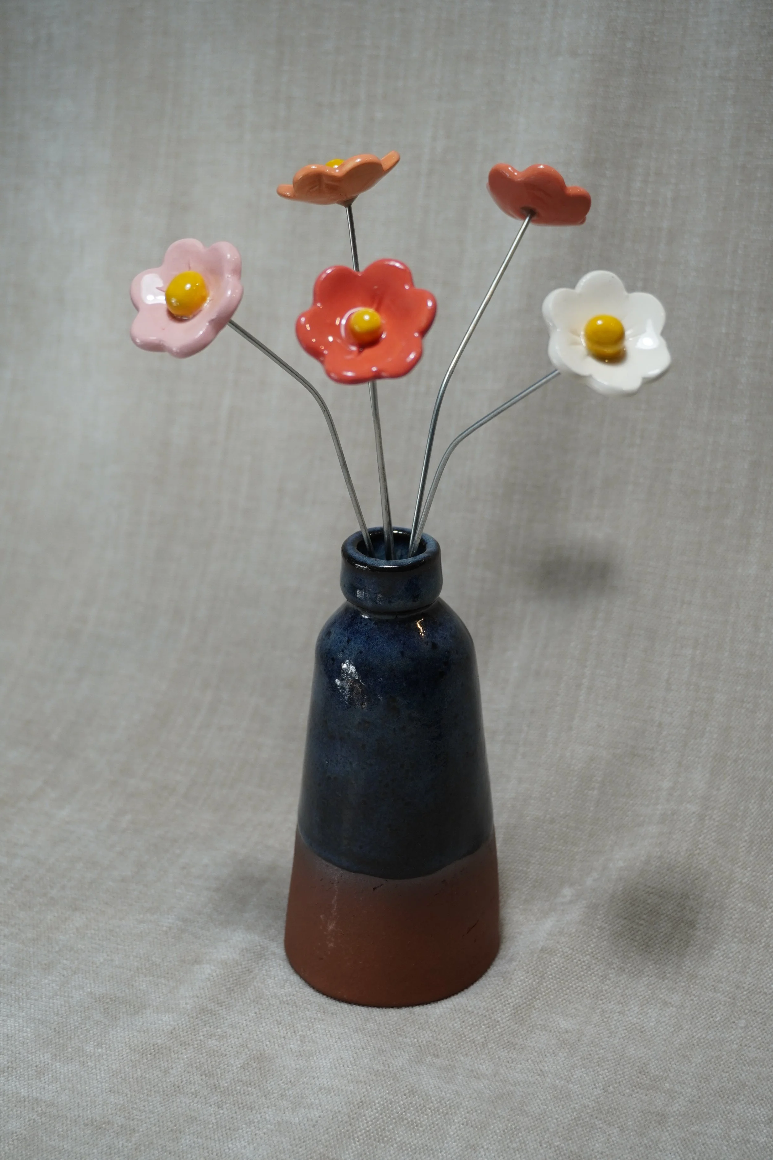 A ceramic pottery vase with a wide, rounded terracotta base, narrow neck, and a blue glaze, holding five ceramic flowers in pink, white, orange, yellow and red with metal stems, displayed on a light neutral background.