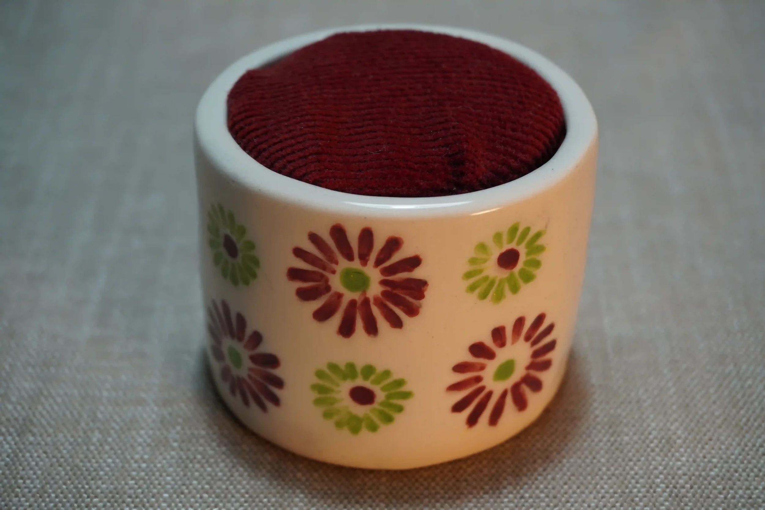 A ceramic pincushion with a red and green firework pattern , filled with red velvet cushion. The pincushion is shown on a textured surface.