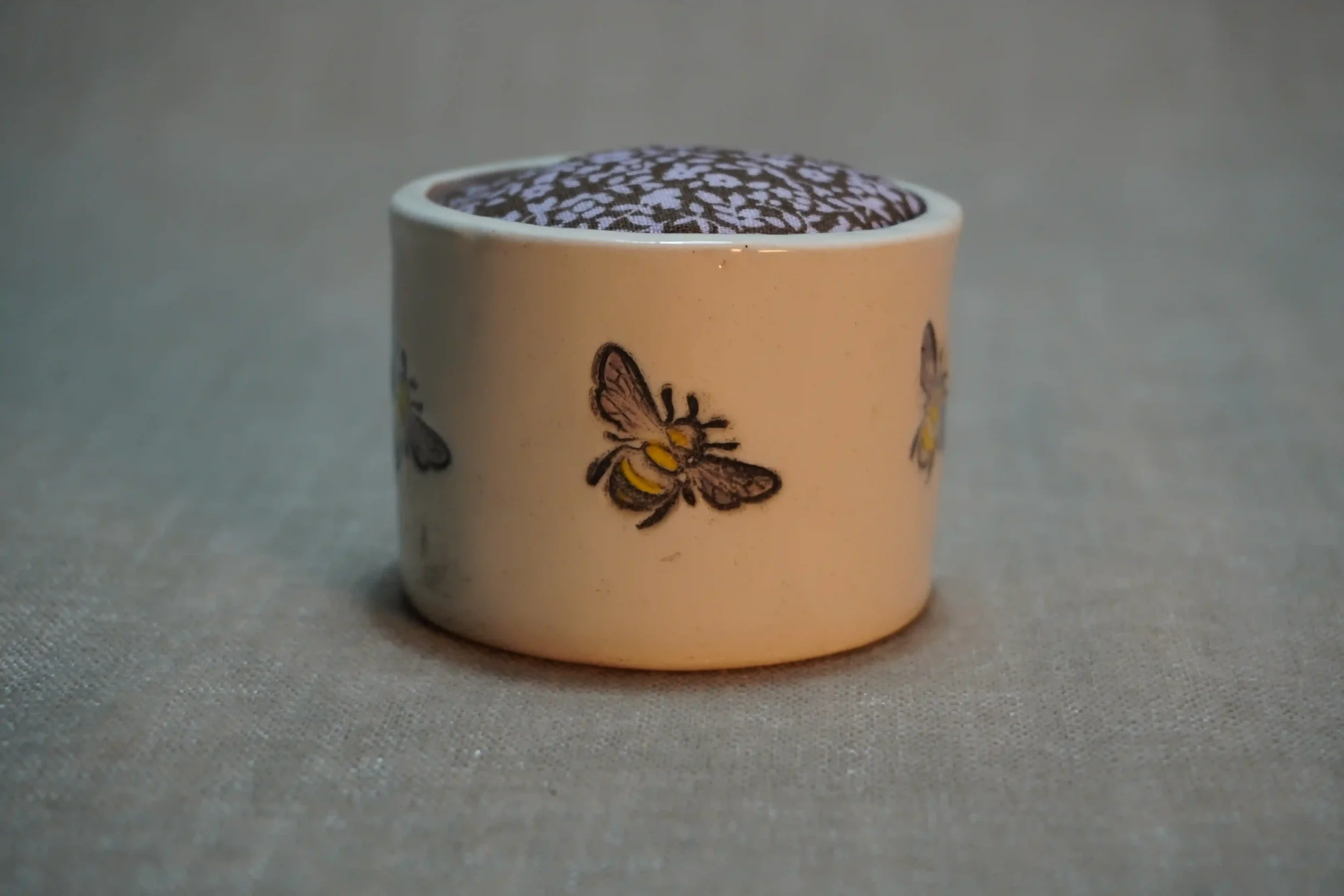 A ceramic pincushion with bee design, filled with a brown and pink floral patterned cushion. The pincushion is shown on a textured surface