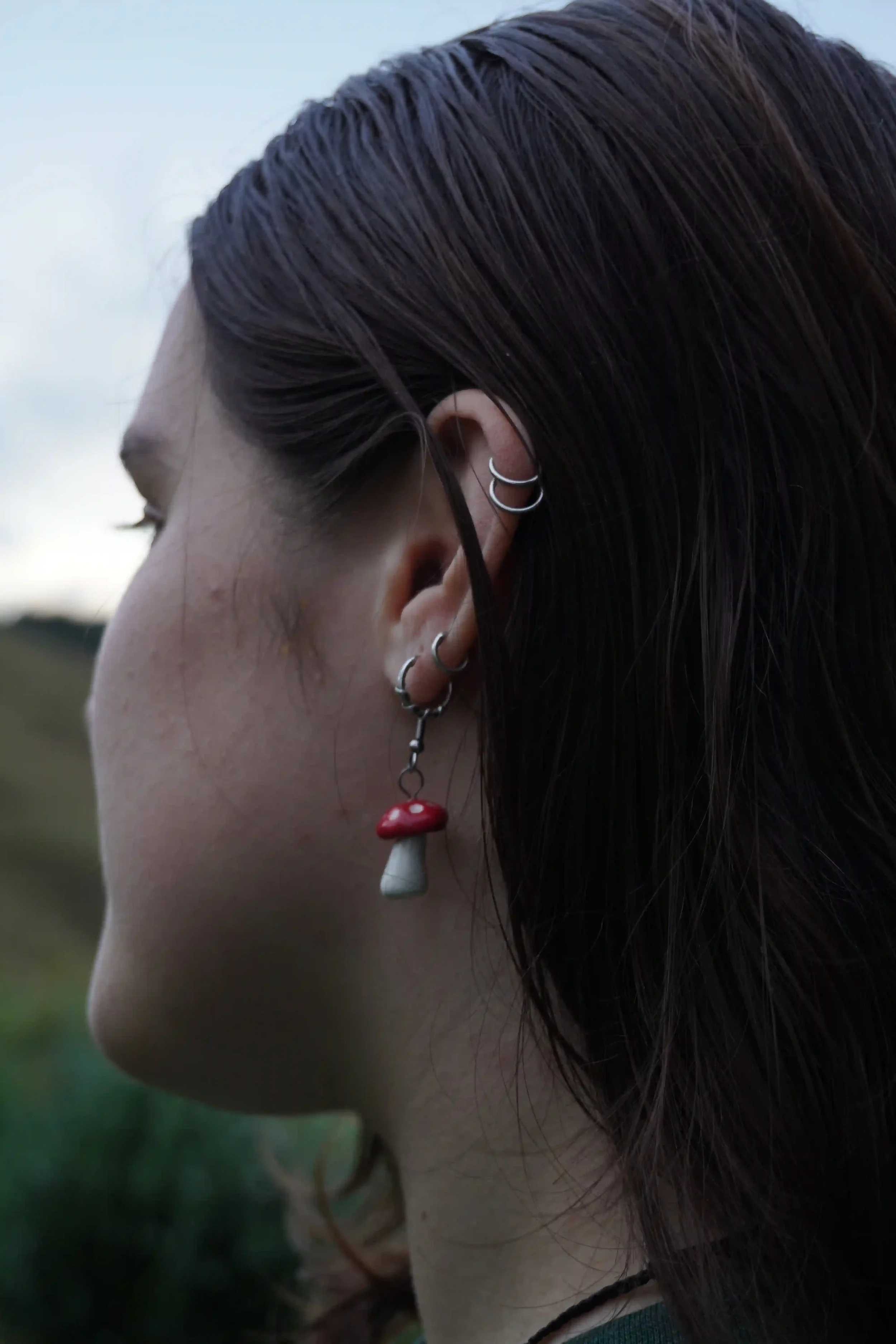 Close-up of a woman's head showing dark brown hair styled in a sleek, wet look. She has multiple silver earrings in her ear, including a hook and hoop design, with a small mushroom-shaped charm hanging from one of the hoops. The background is blurred outdoor scenery with greenery.