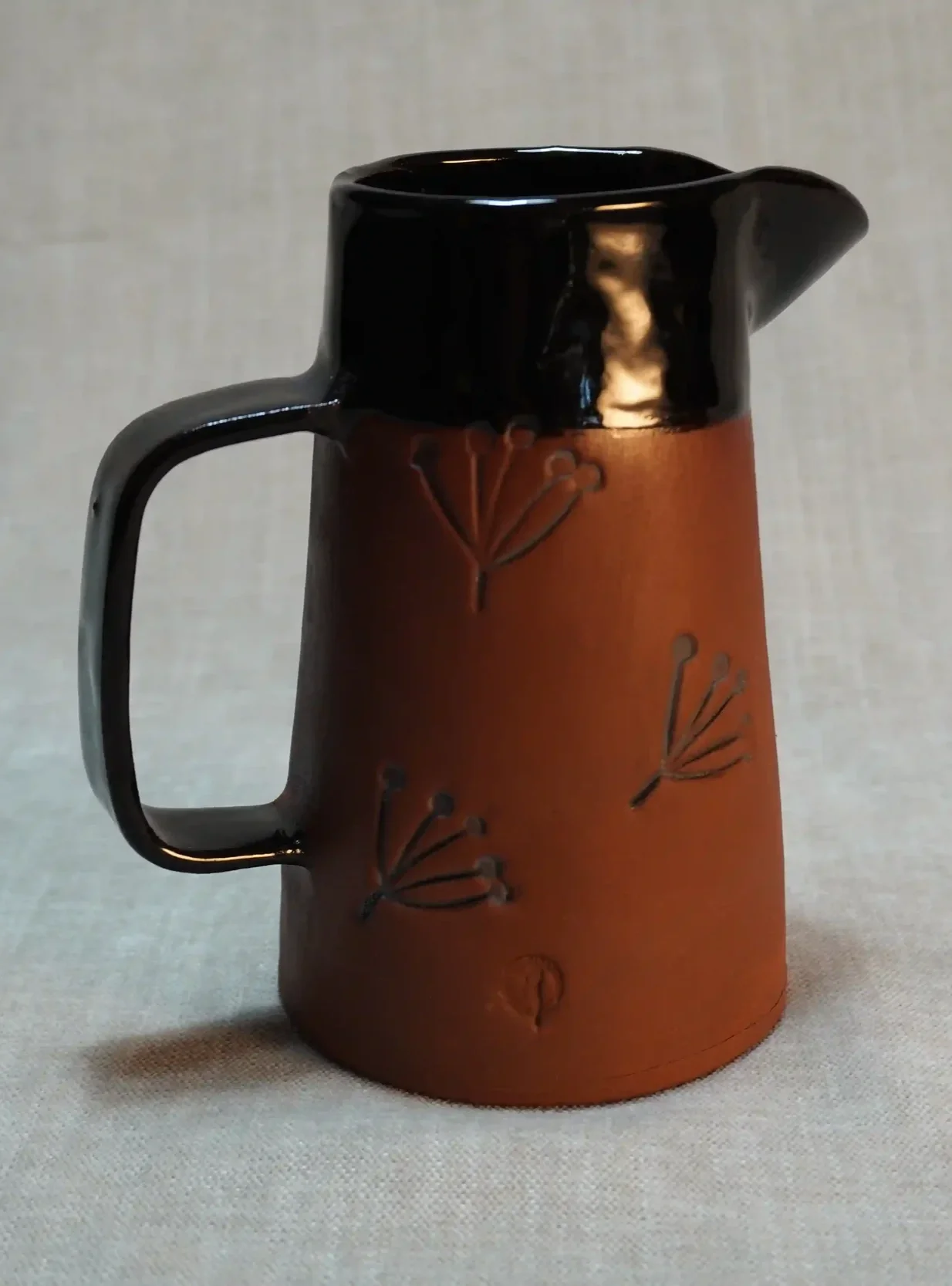 A ceramic mug with a black top and handle, and a brown body decorated with carved dandelion flower motifs.