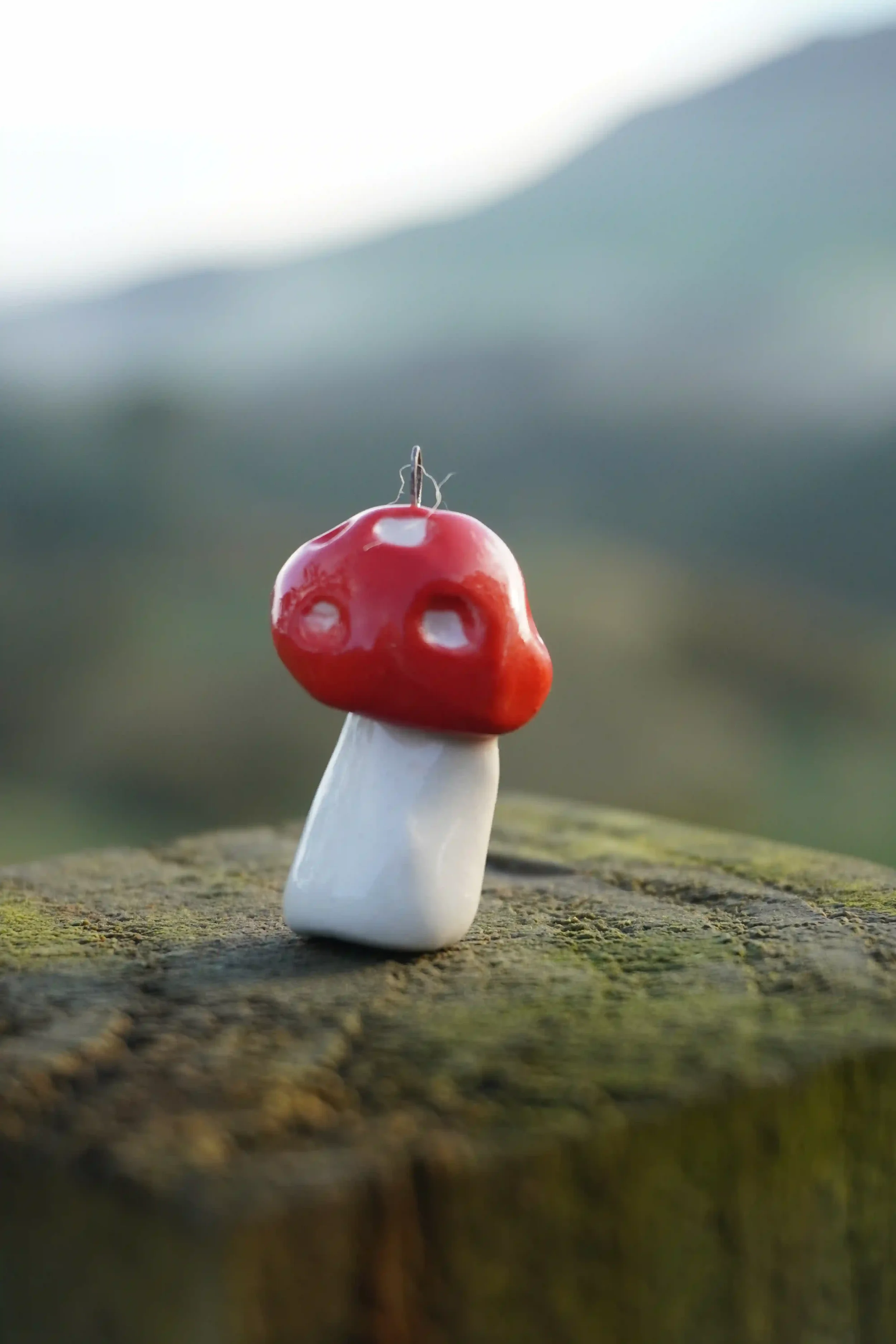 Close-up of a handmade ceramic mushroom with a red cap and white stem displayed on a rough, mossy wooden surface, blurred natural background.
