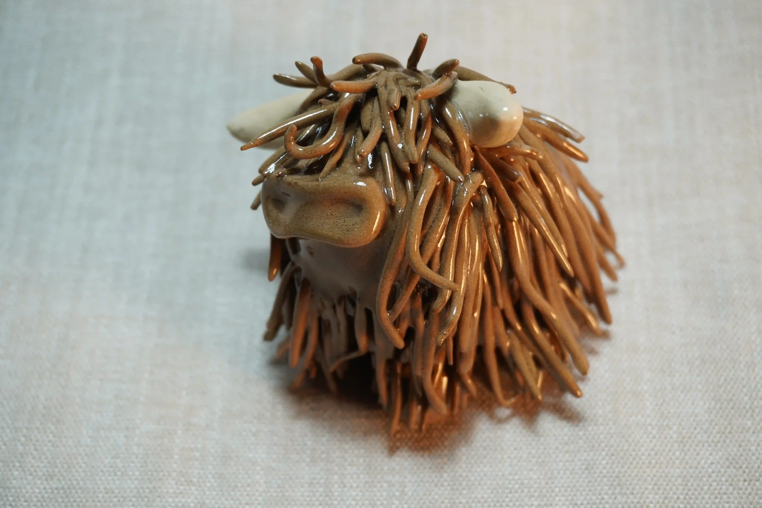 Frontal view of a handmade pottery ceramic sculpture of a Highland Cow ornament with milk chocolate-coloured hair and beige horns.