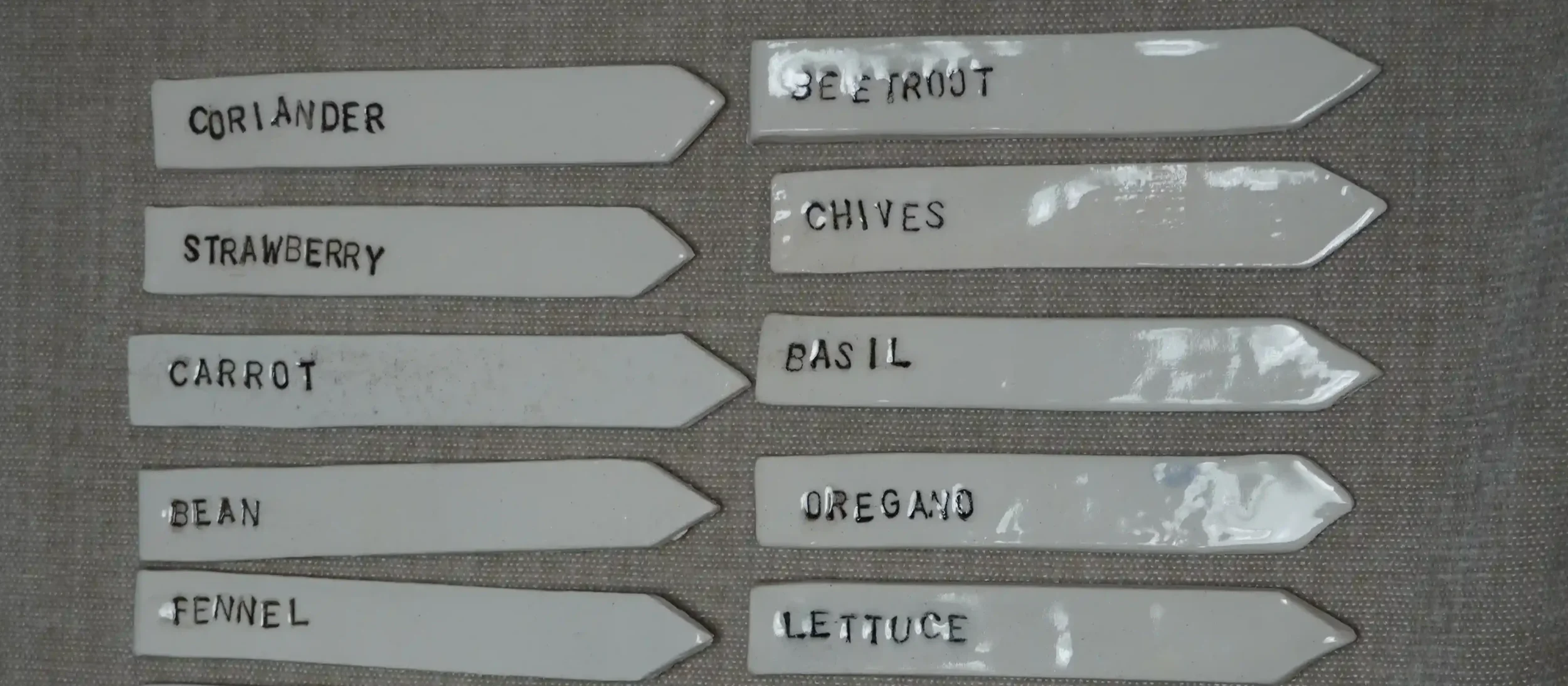 White ceramic plant labels with black handwritten text, arranged in two columns, indicating different herbs such as coriander, strawberry, carrot, bean, fennel, beetroot, chives, basil, oregano, and lettuce.
