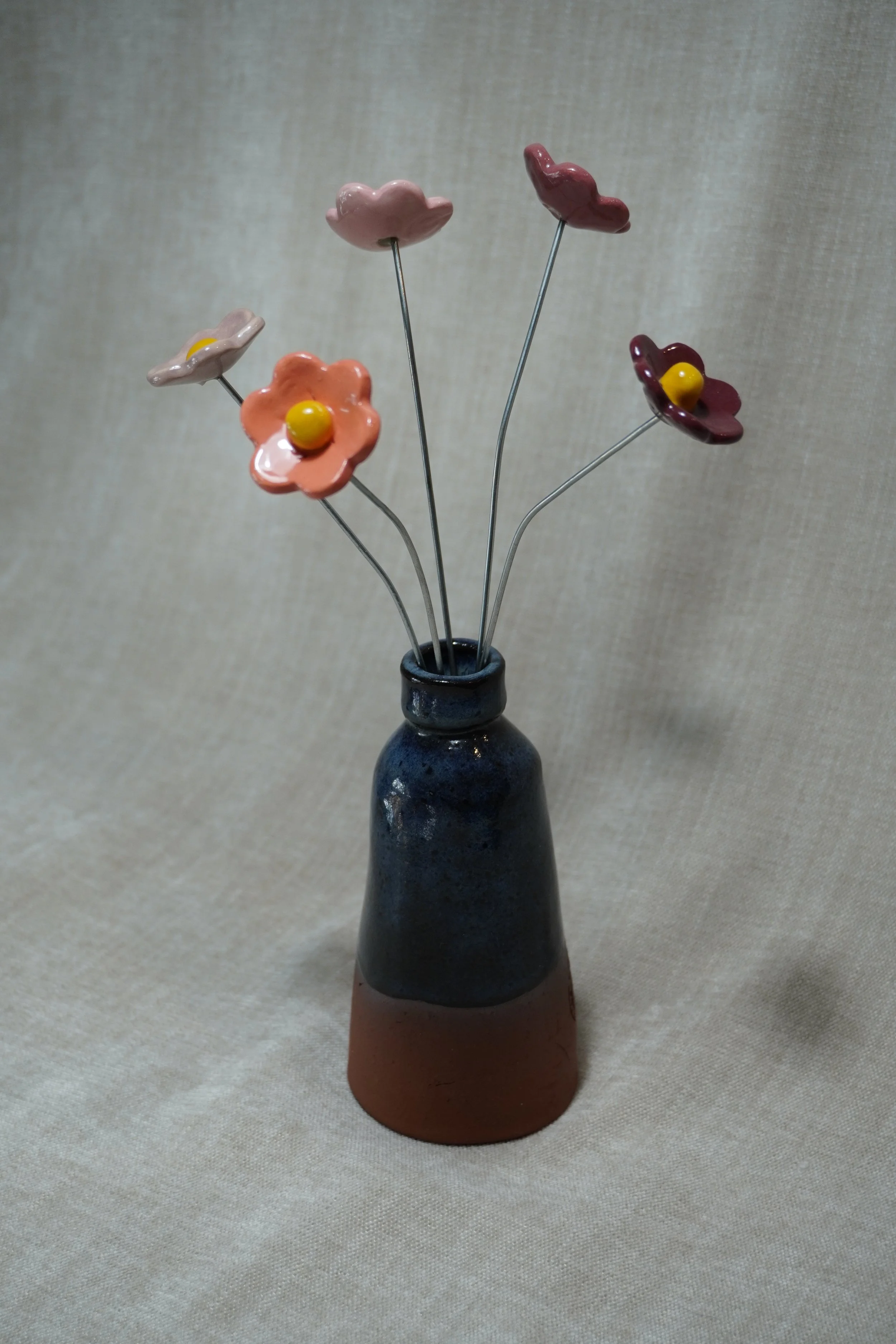 A ceramic pottery vase with a wide, rounded terracotta base, narrow neck, and a blue glaze, holding five ceramic purple, pink, and orange flowers with metal stems, displayed on a light neutral background.