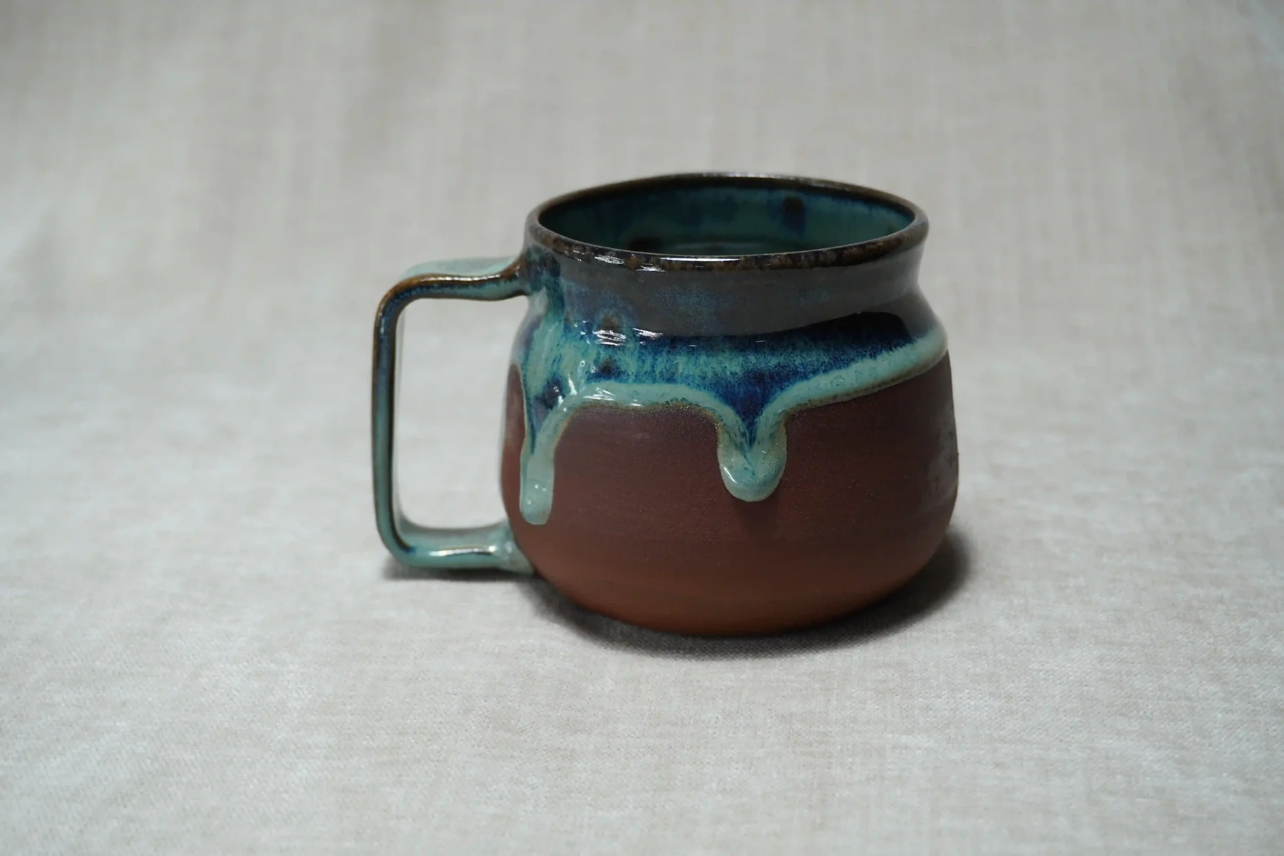 A ceramic mug with a handle, featuring a blue-green glaze, sitting on a plain, light-colored surface.