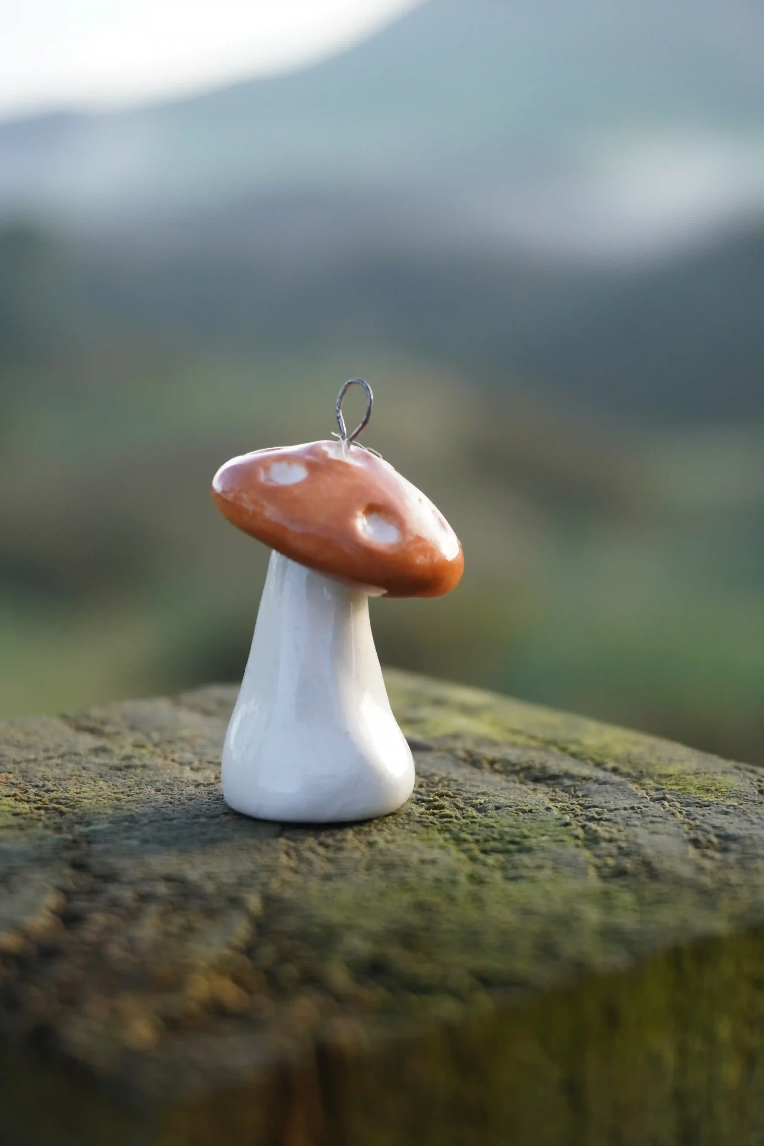 Close-up of a handmade ceramic mushroom with a small metal hook on top, placed on a rough wooden surface outdoors.