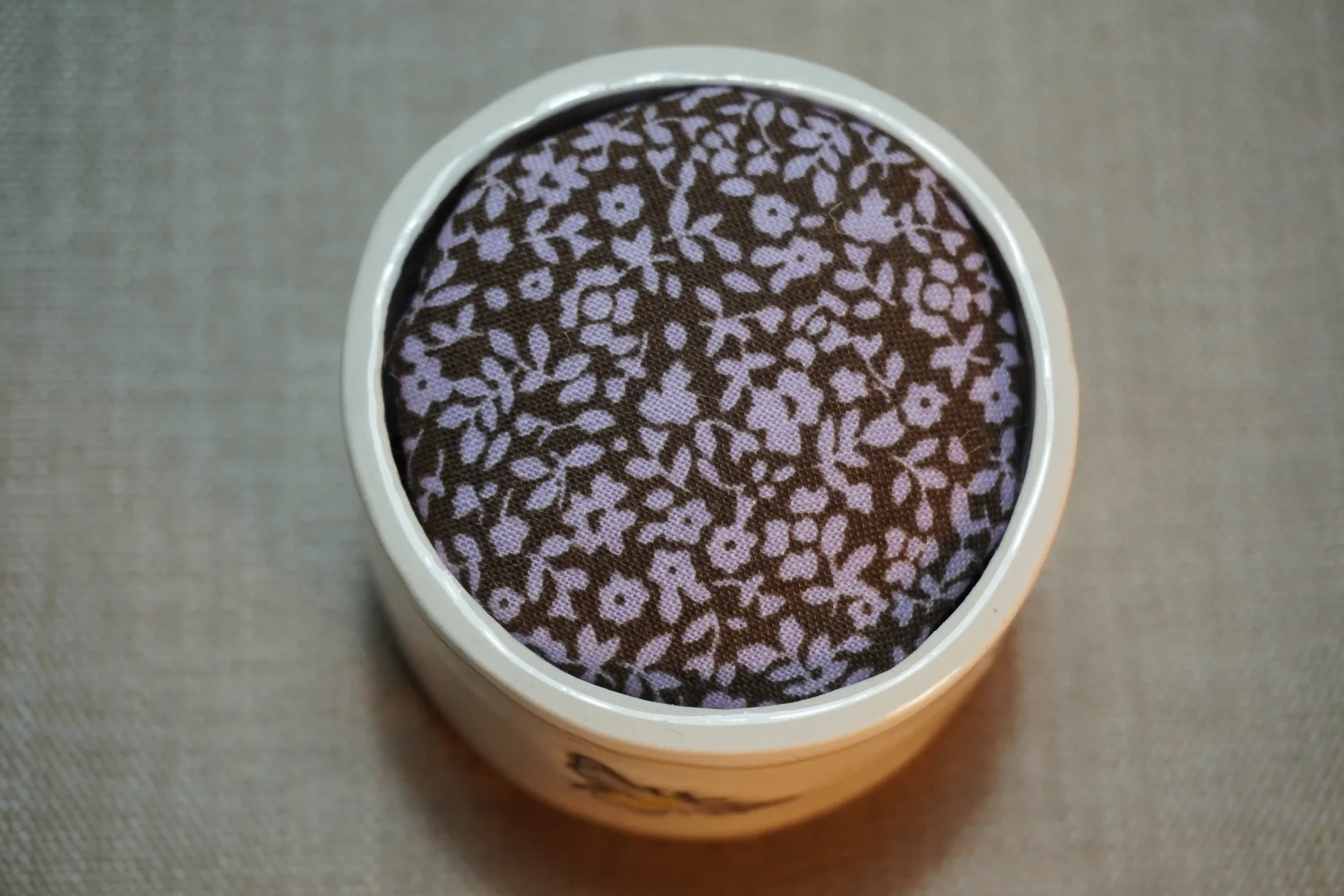 View from above of a ceramic pincushion filled with a brown and pink floral patterned cushion. The pincushion is shown on a textured surface.