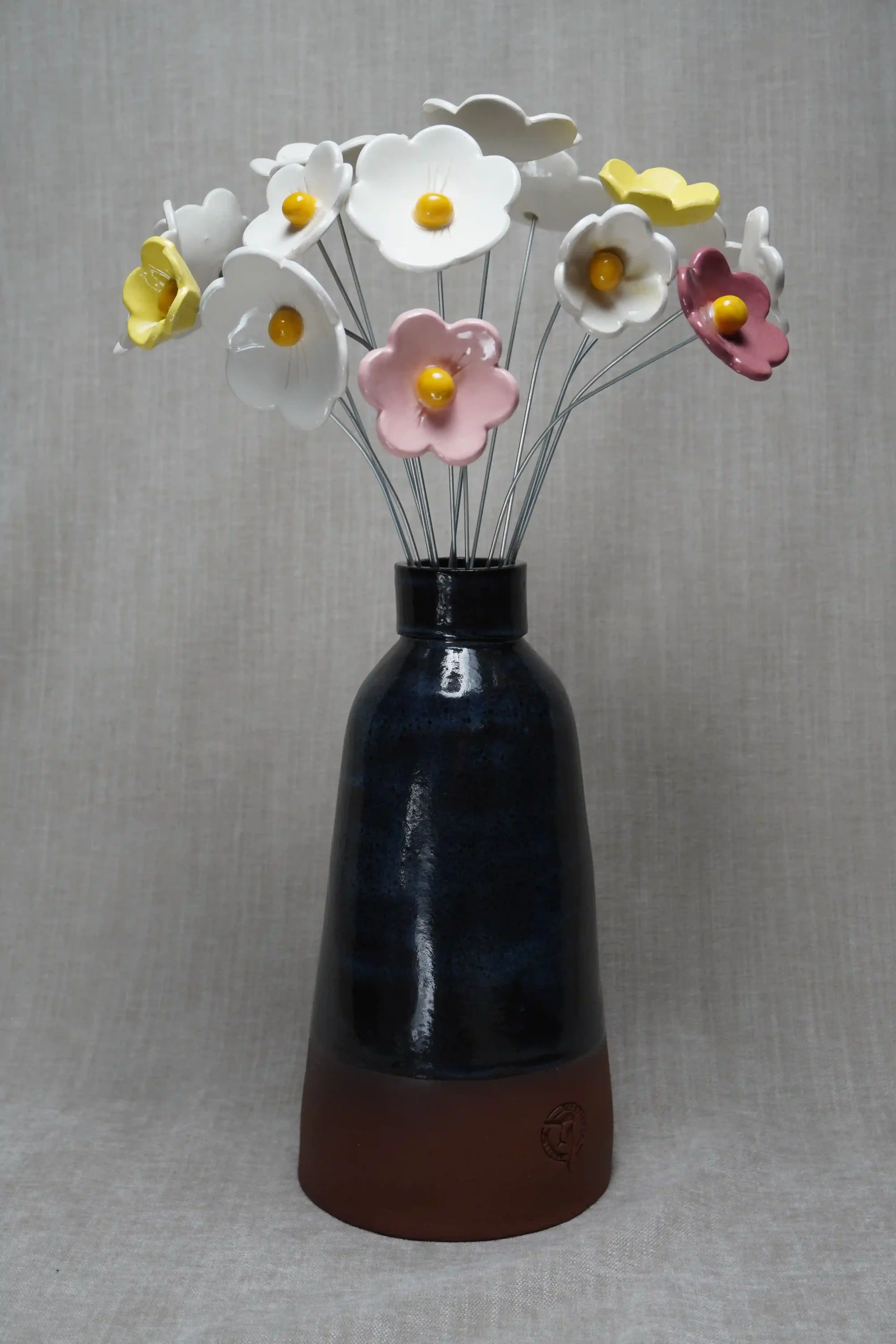 Decorative flower arrangement with white, yellow and pink ceramic flowers in pastel colors in a blue-glazed ceramic vase that has a terracotta brown base.