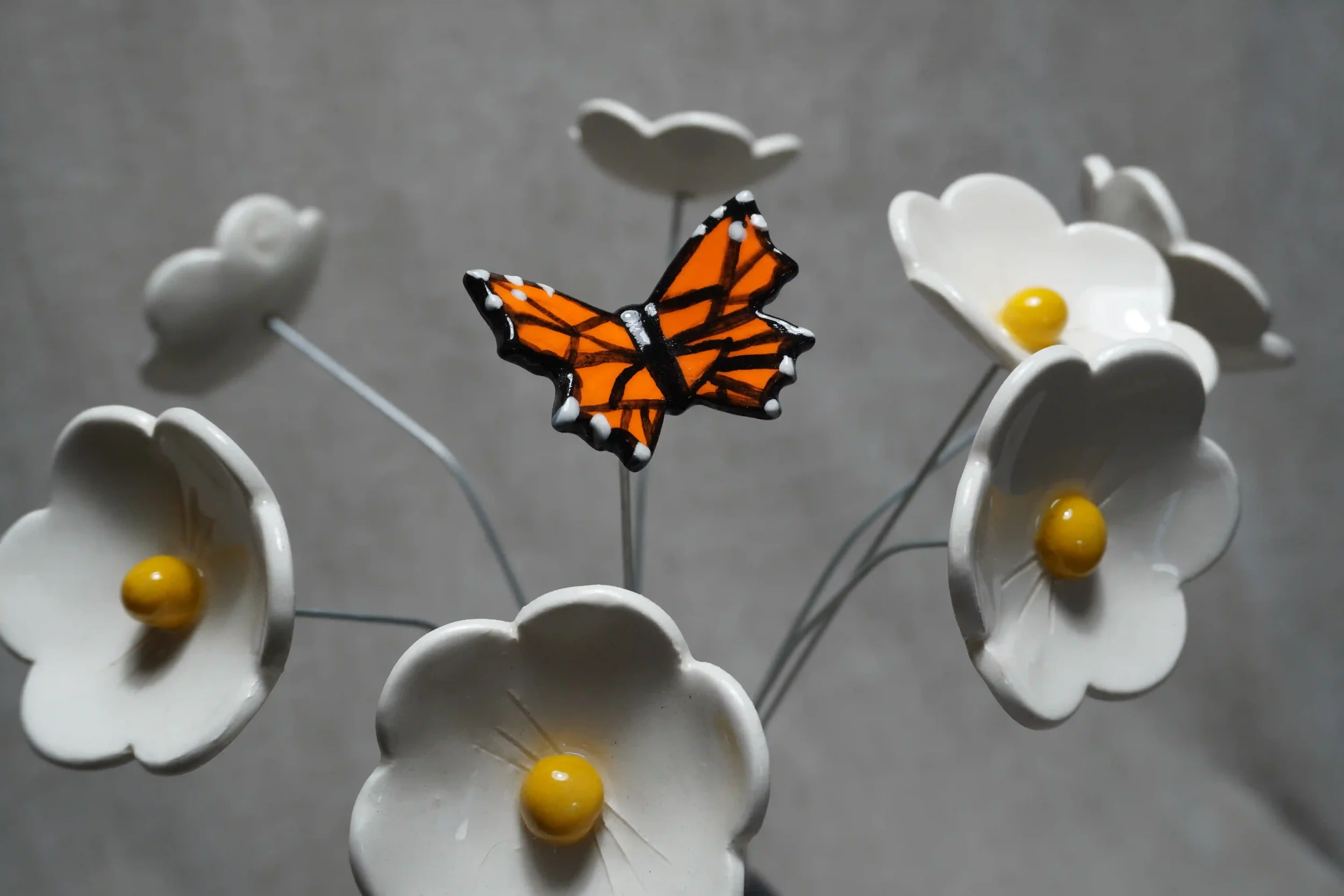 An orange and black ceramic monarch butterfly on a metal stem displayed flying among white daisy flowers with yellow centres.
