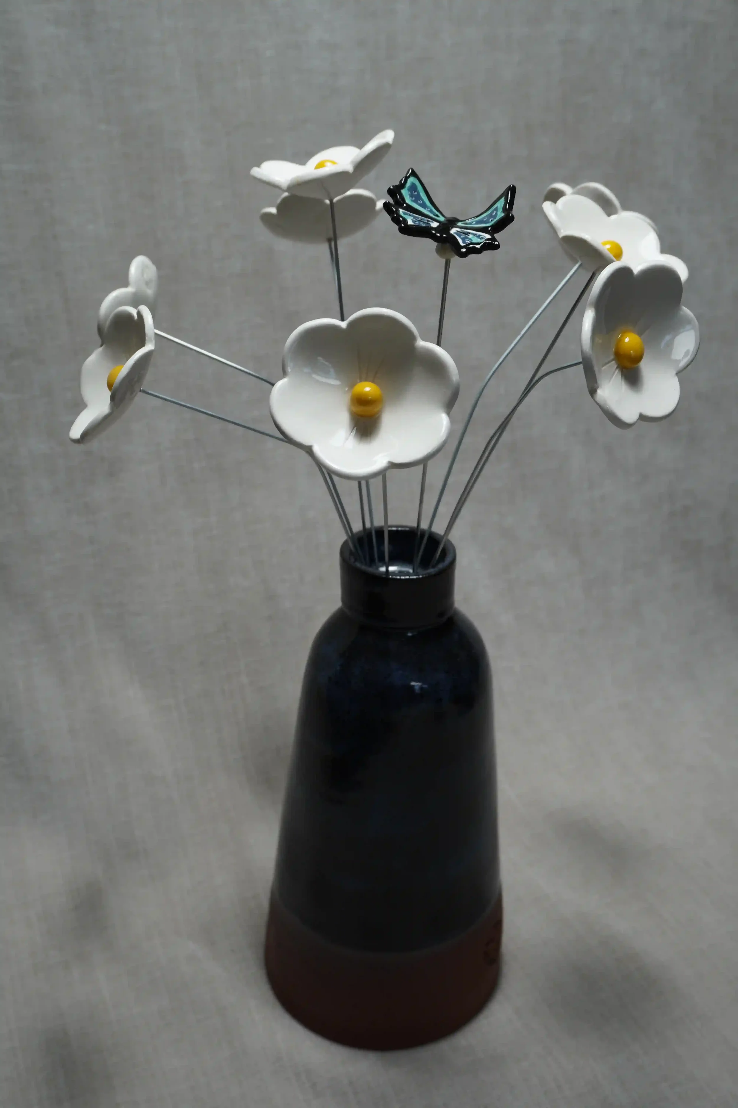 A dark blue-glazed handmade ceramic vase holding white ceramic Daisy flowers with yellow centers and a decorative ceramic blue butterfly, set against a neutral background.