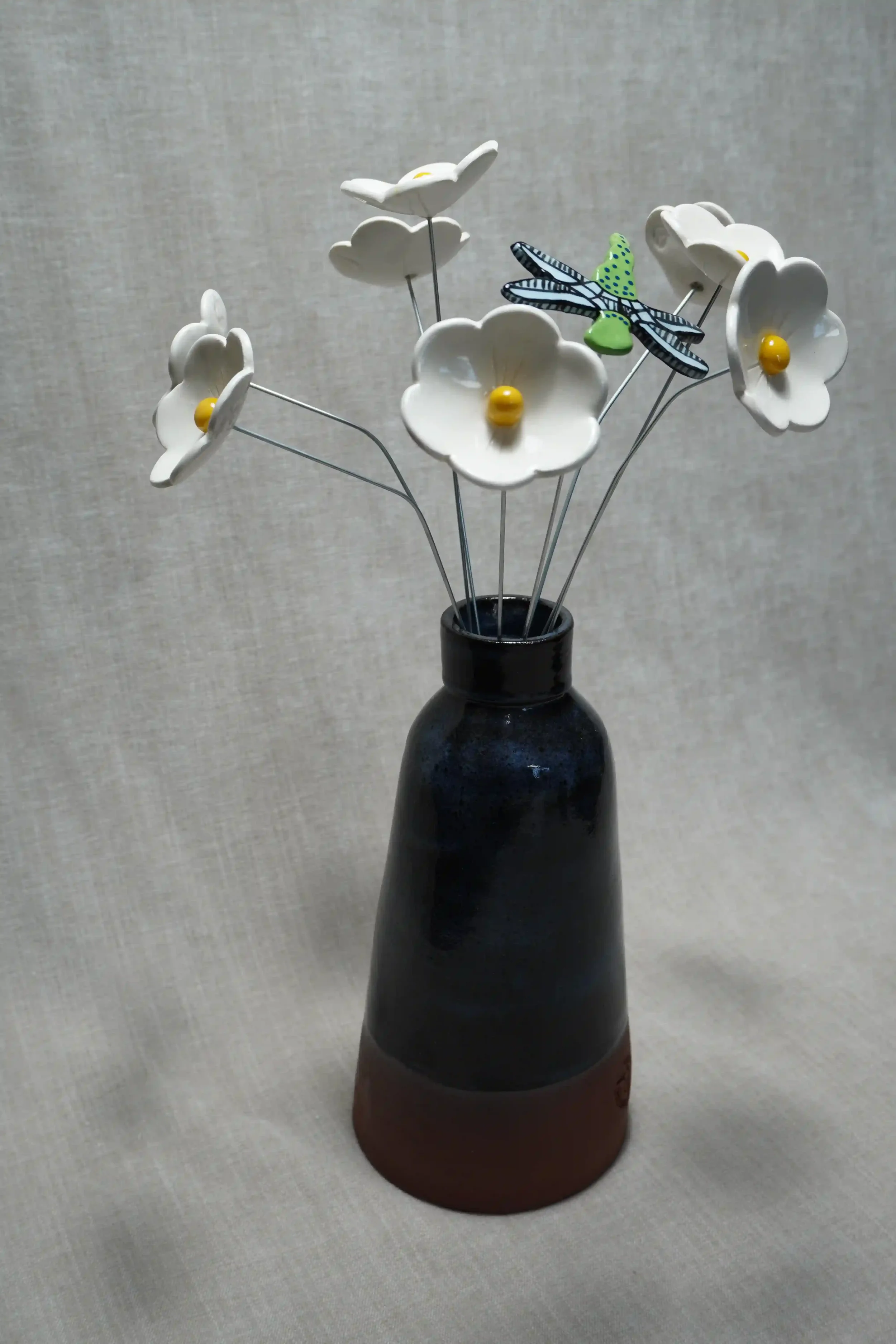 A black and brown ceramic vase holding white ceramic flowers with yellow centers and a decorative dragonfly with with light blue wings and a green body with blue speckles.