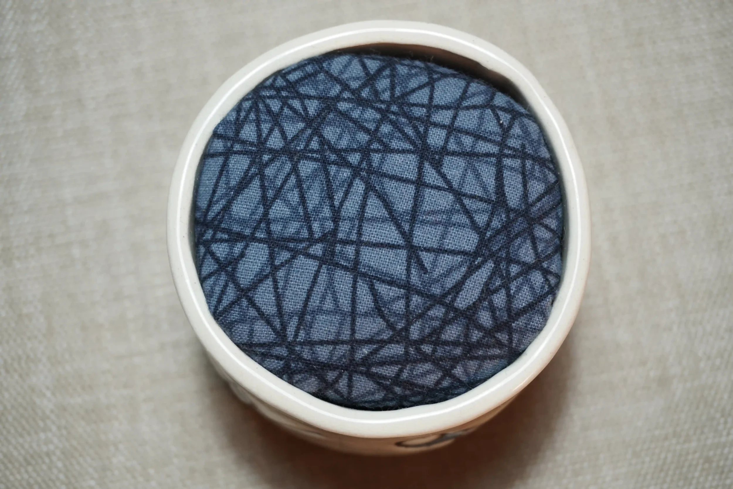 View from above of a ceramic pincushion with a blue patterned fabric-covered cushion, featuring black intersecting lines, shown on a textured surface.