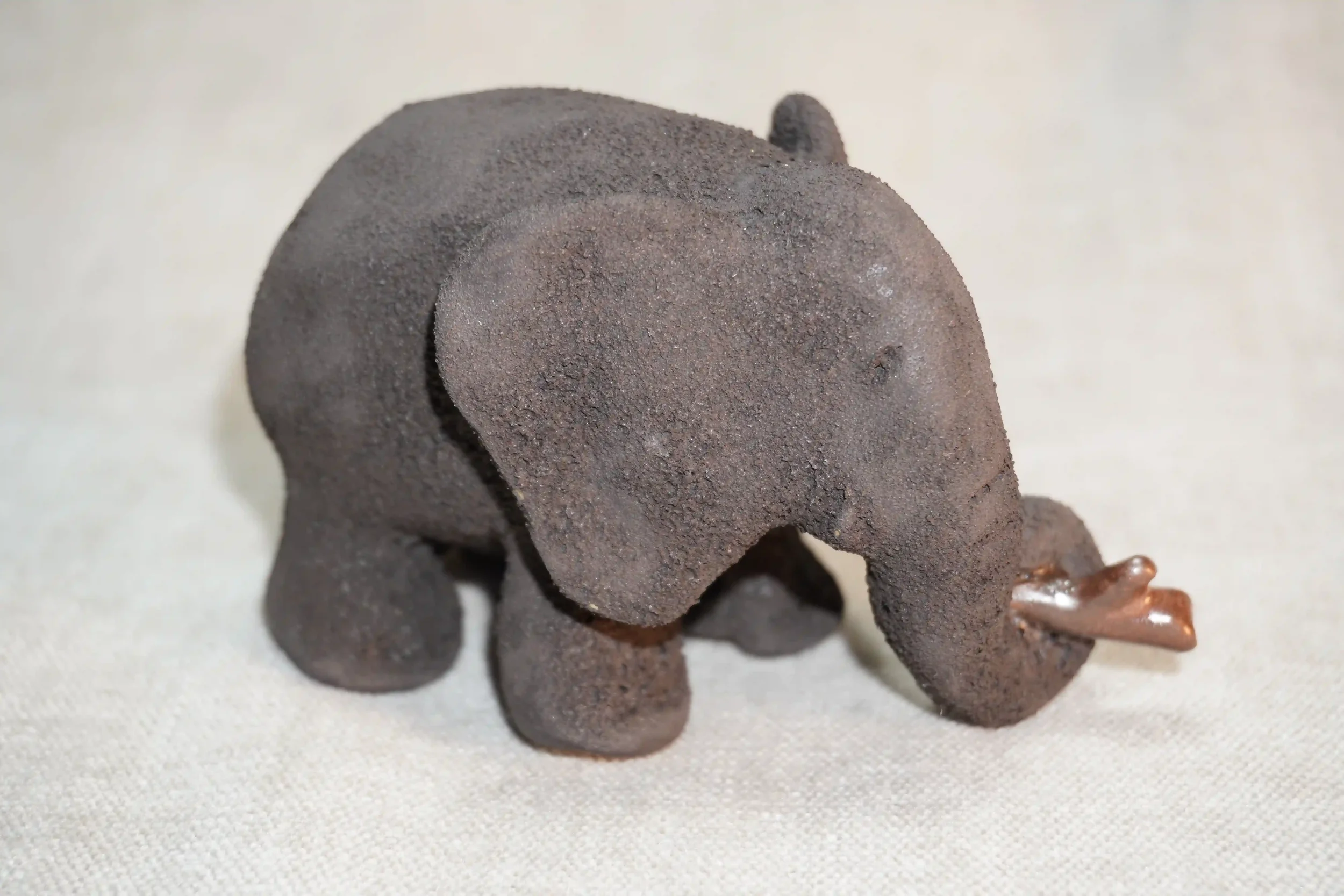 Side view of a handmade pottery elephant figurine with a dark grey textured surface and holding a branch in it's trunk, placed on a light-colored fabric surface.