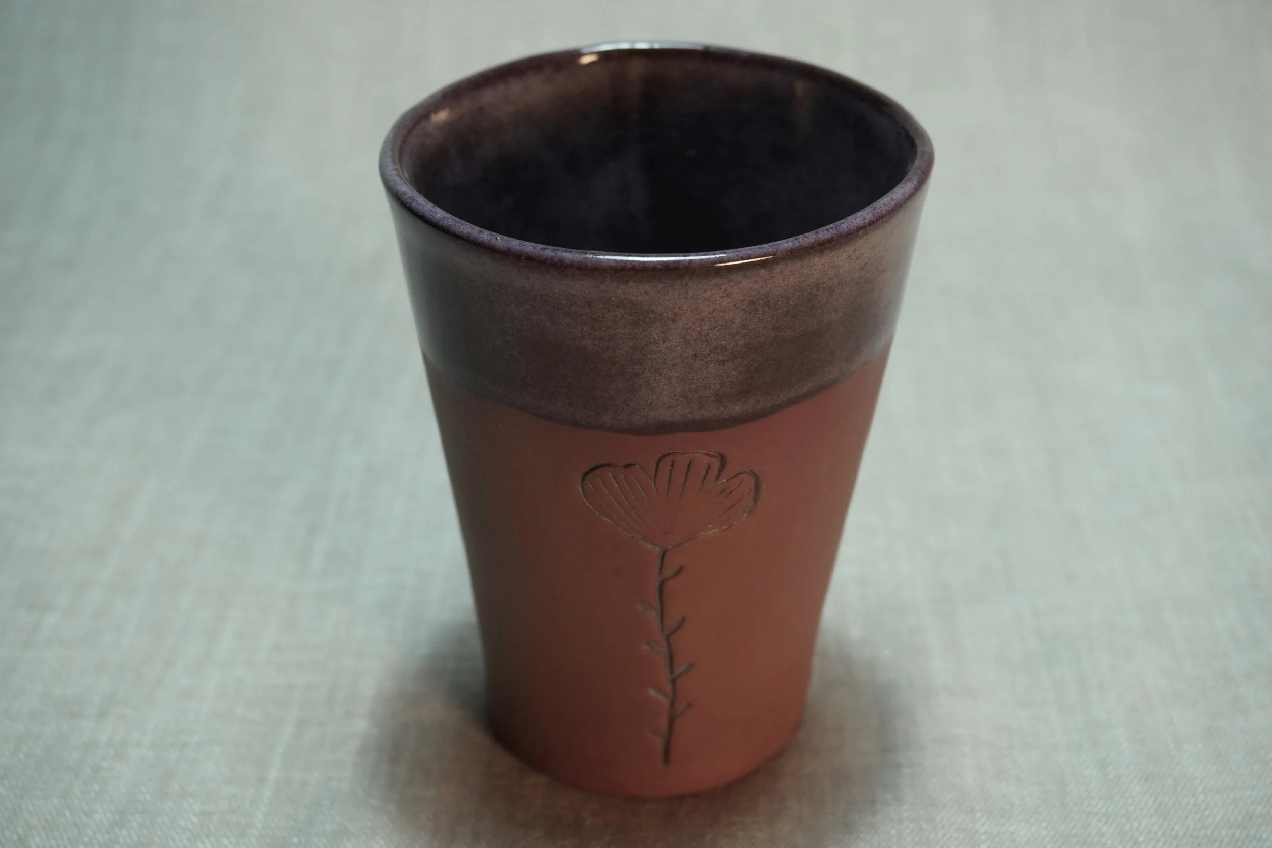 A pottery tumbler with purple glaze, a terracotta brown base, decorated with a flower with a stem and leaves on the outside. Shown displayed on a light textured surface.