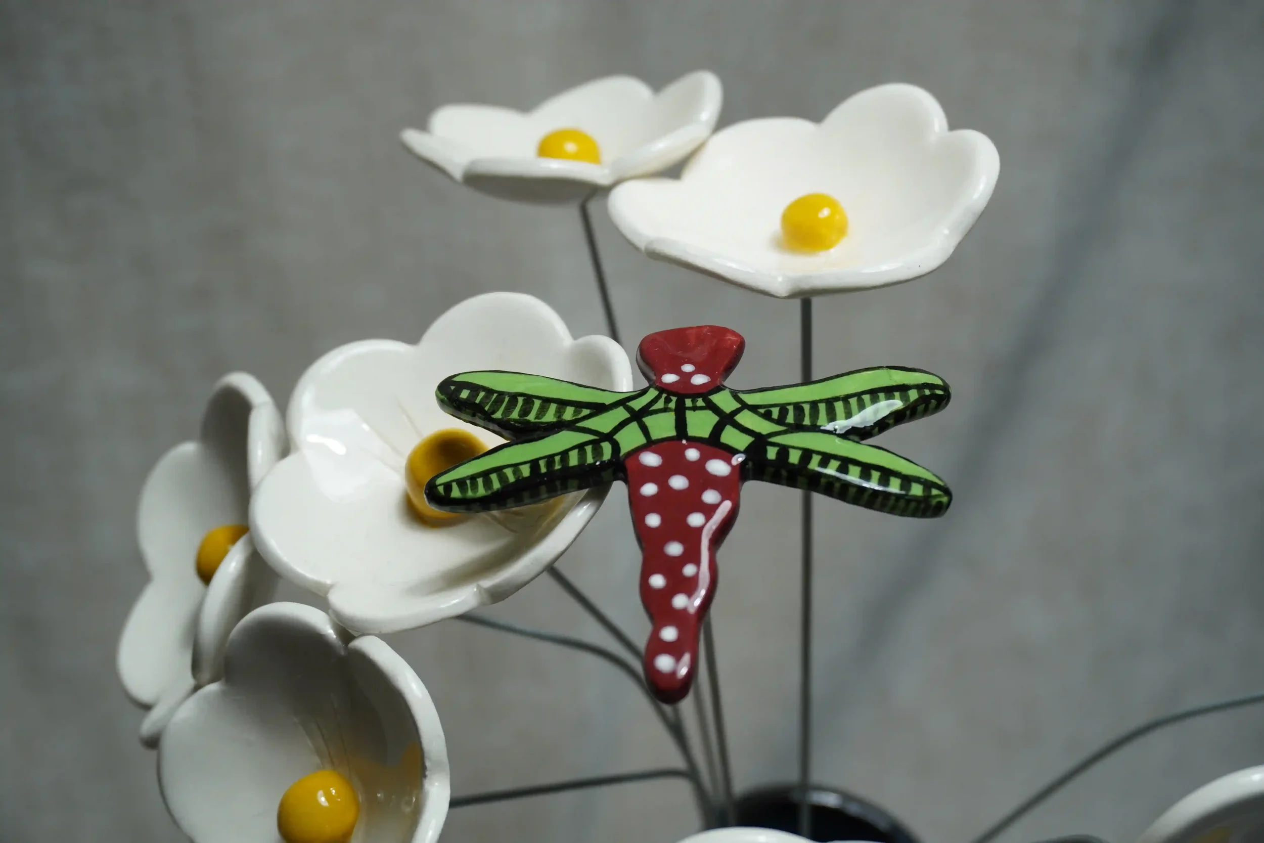 Ceramic dragonfly ornament with a red and white speckled body and green and black wings, on a metal stem. Shown displayed among white Daisies with yellow centres.