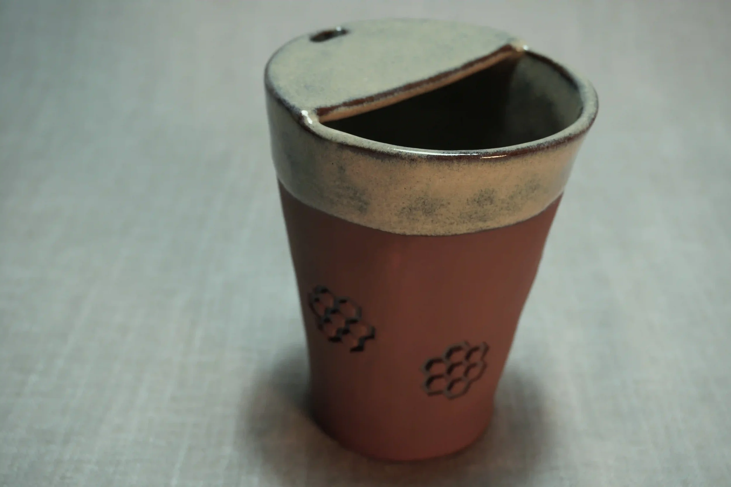 A ceramic travel mug with a terracotta brown base, a cream-colored top rim, and a decorative honeycomb pattern on the outside, placed on a light surface.
