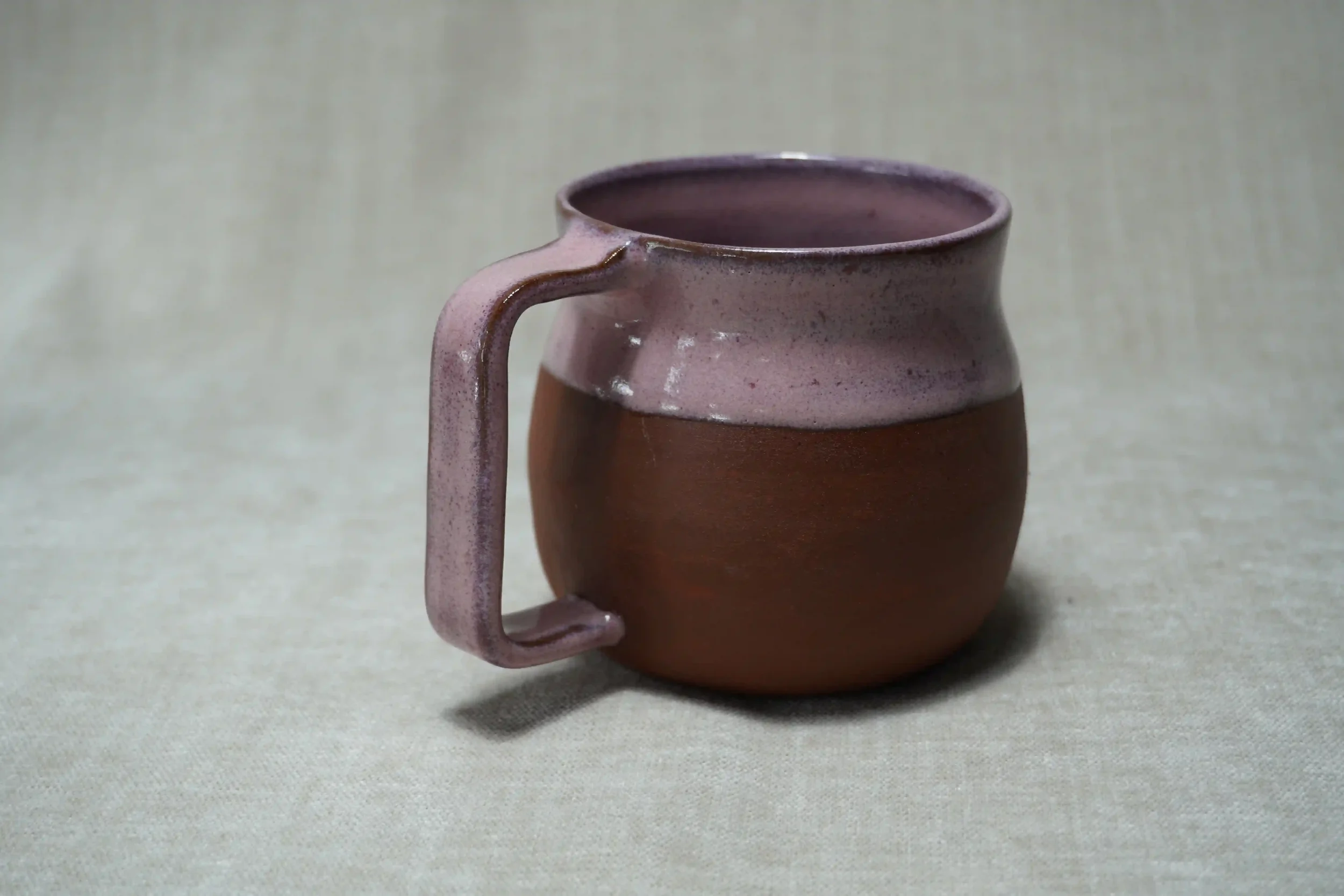 A ceramic pottery belly mug with a pink glaze on its handle and top, and an unglazed terracotta brown body, placed on a beige surface with a plain, light-colored background.