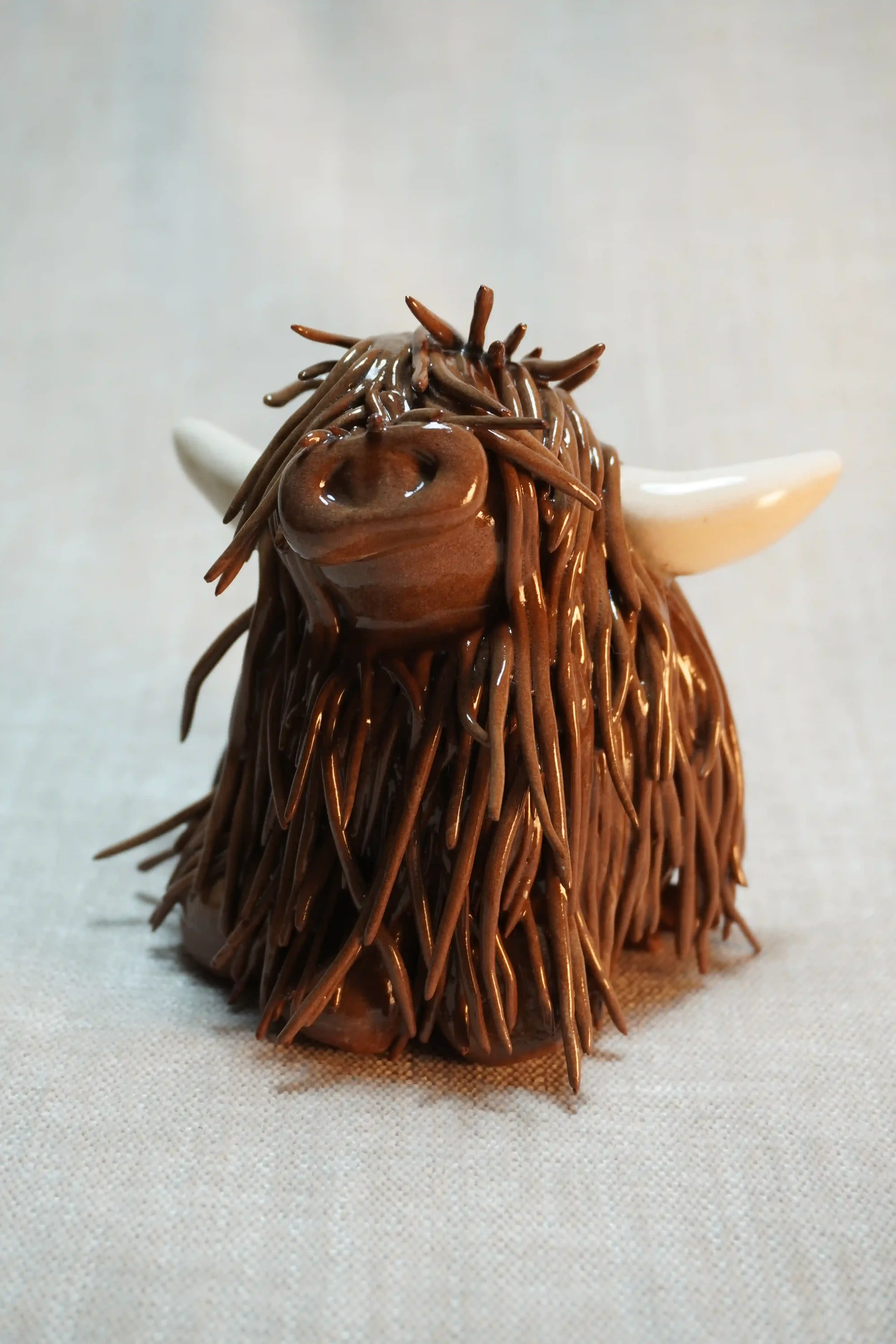 Front view of a handmade ceramic sculpture of a Highland Cow ornament with rich dark brown-coloured hair and beige horns.