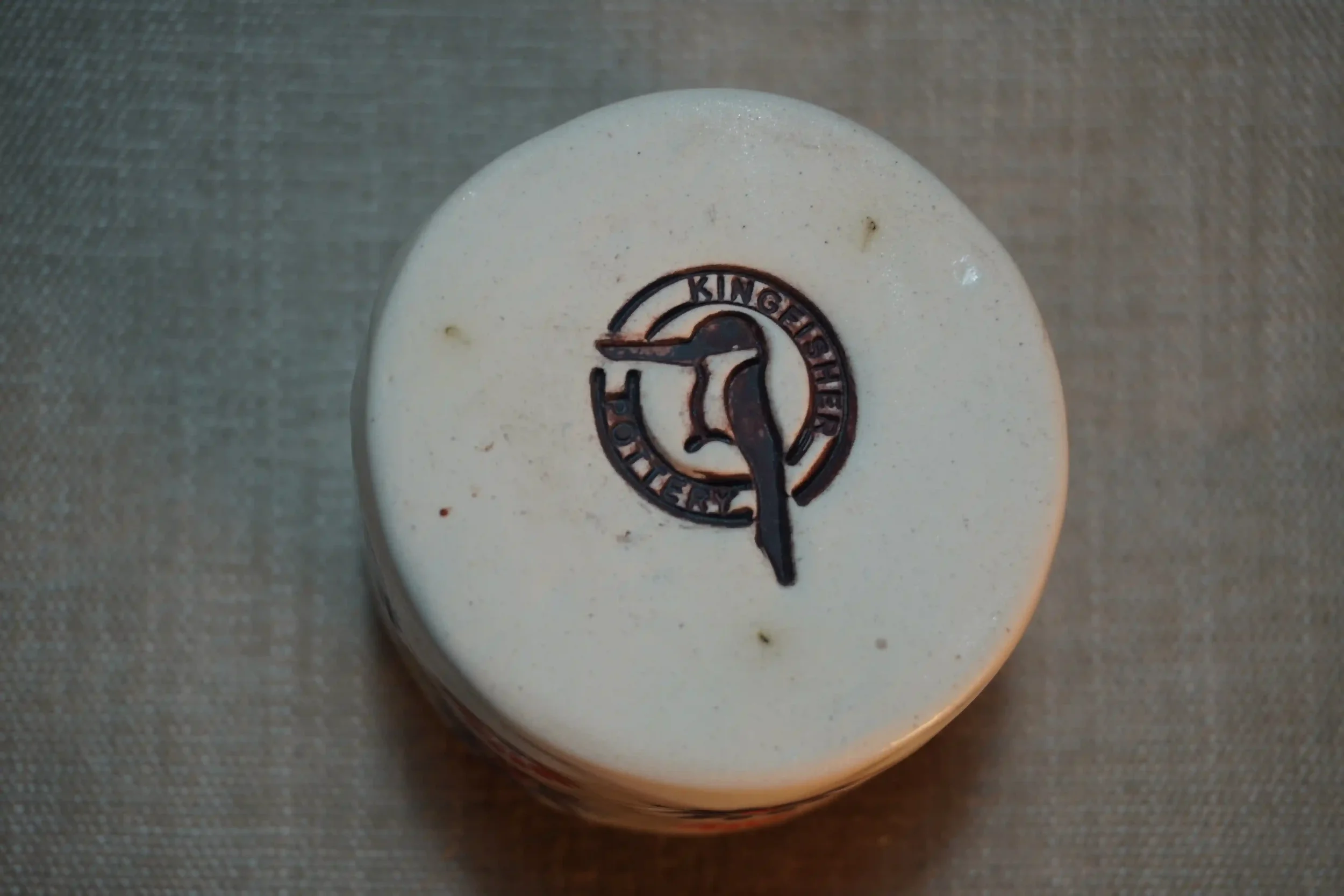 View of a pottery maker's mark on the bottom of a ceramic pincushion. The maker's mark shows a kingfisher and words saying Kingfisher Pottery.