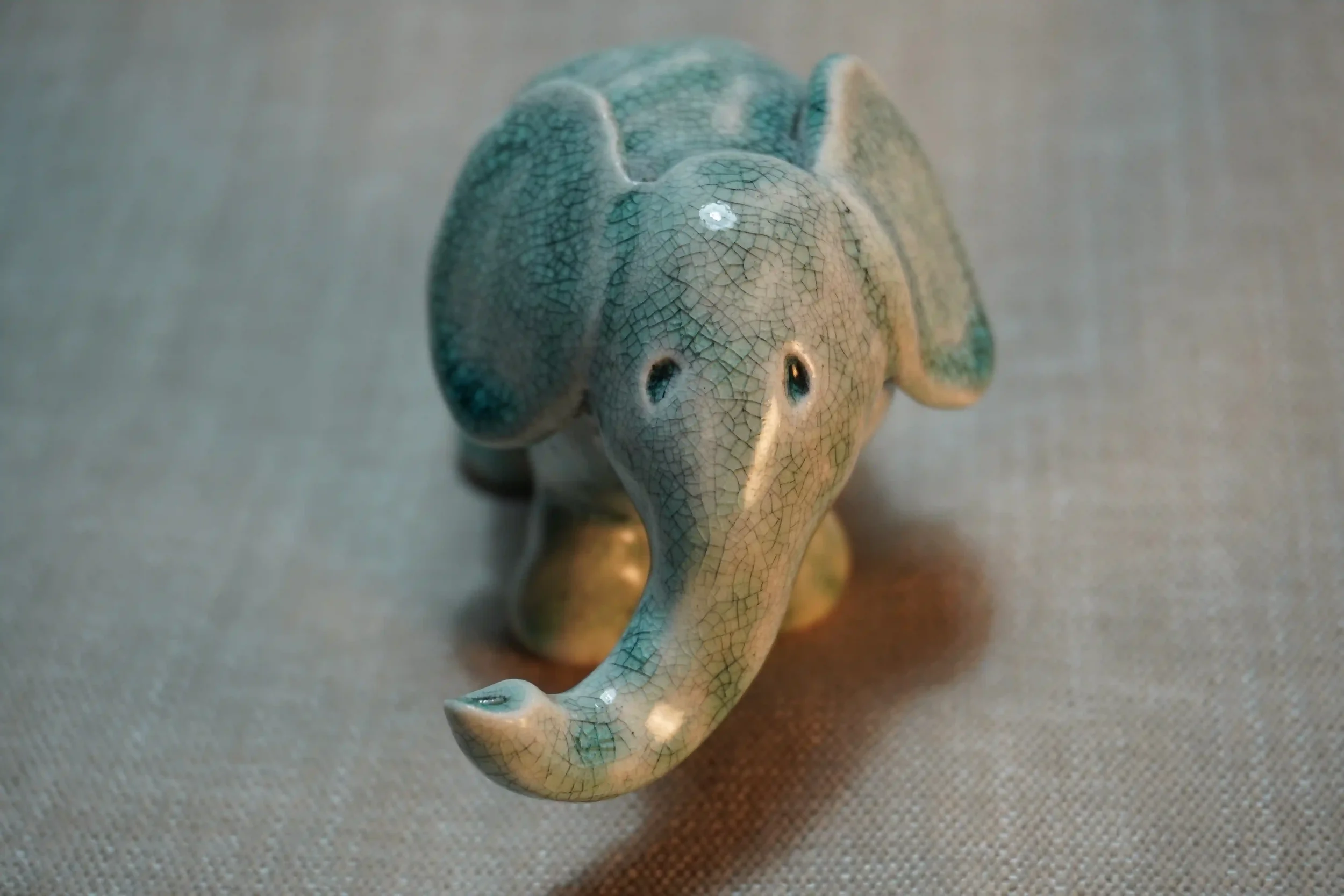 A ceramic figurine of an elephant with a crackled glaze finish, positioned on a neutral-colored fabric surface.